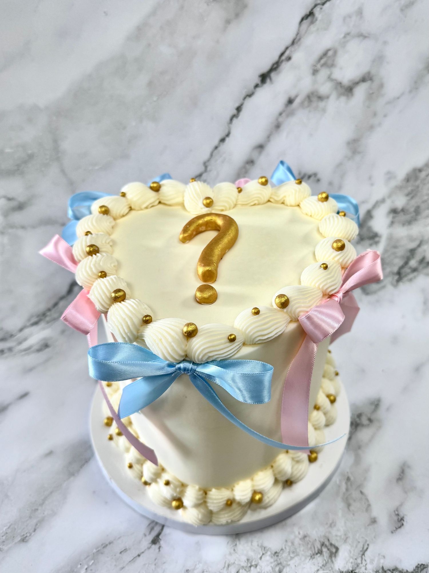 A white cake with gold pearl decorations and pink and blue ribbon accents sits on a marble countertop.