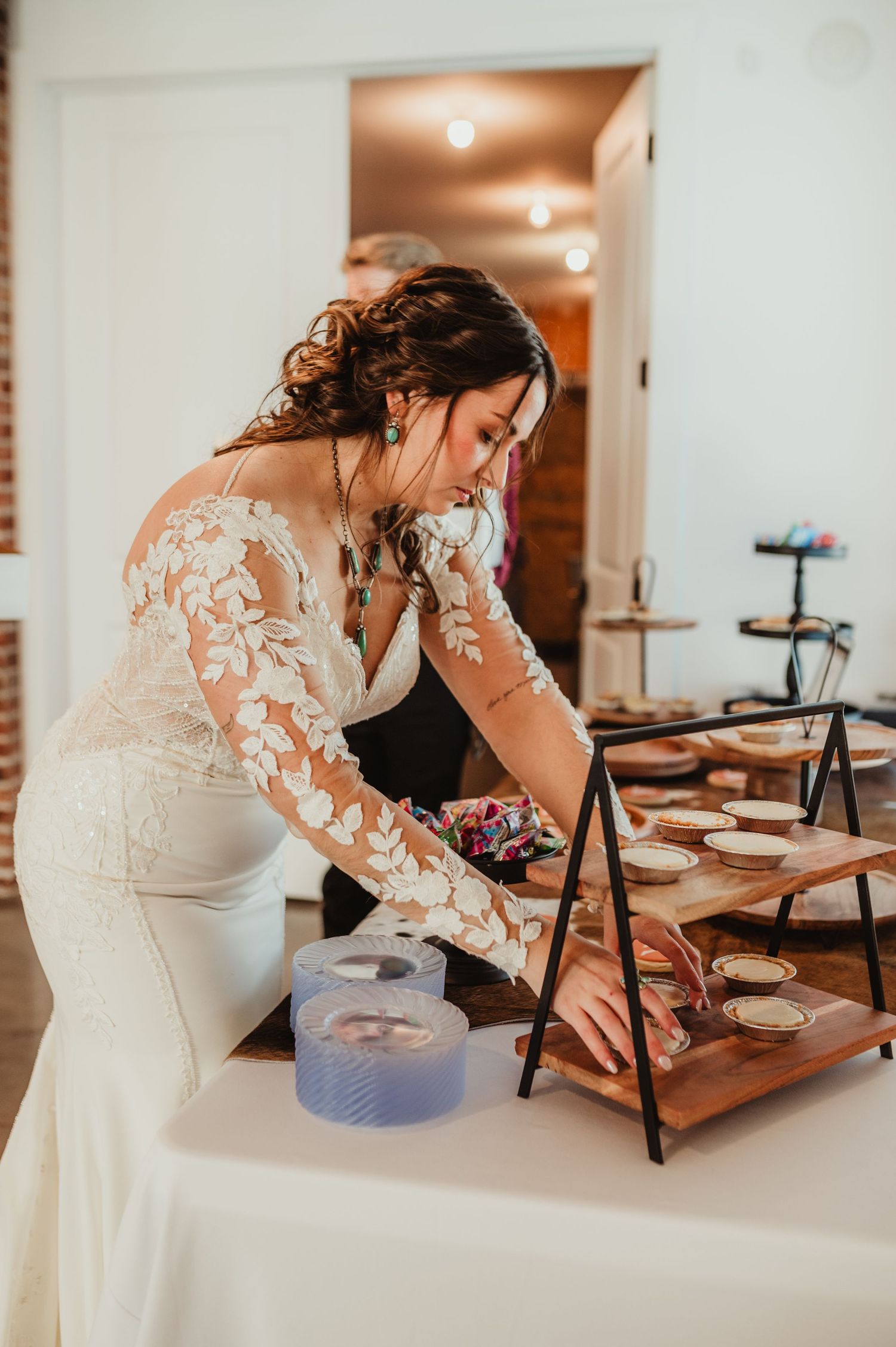 Person in white lace dress arranging desserts on wooden display stands.