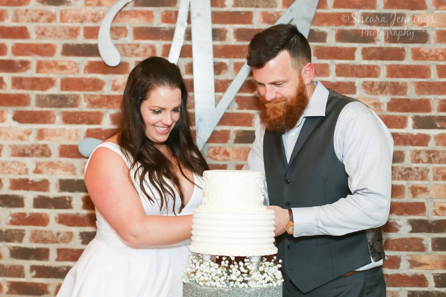 Wedding couple cutting their white tiered cake against a rustic brick wall backdrop.