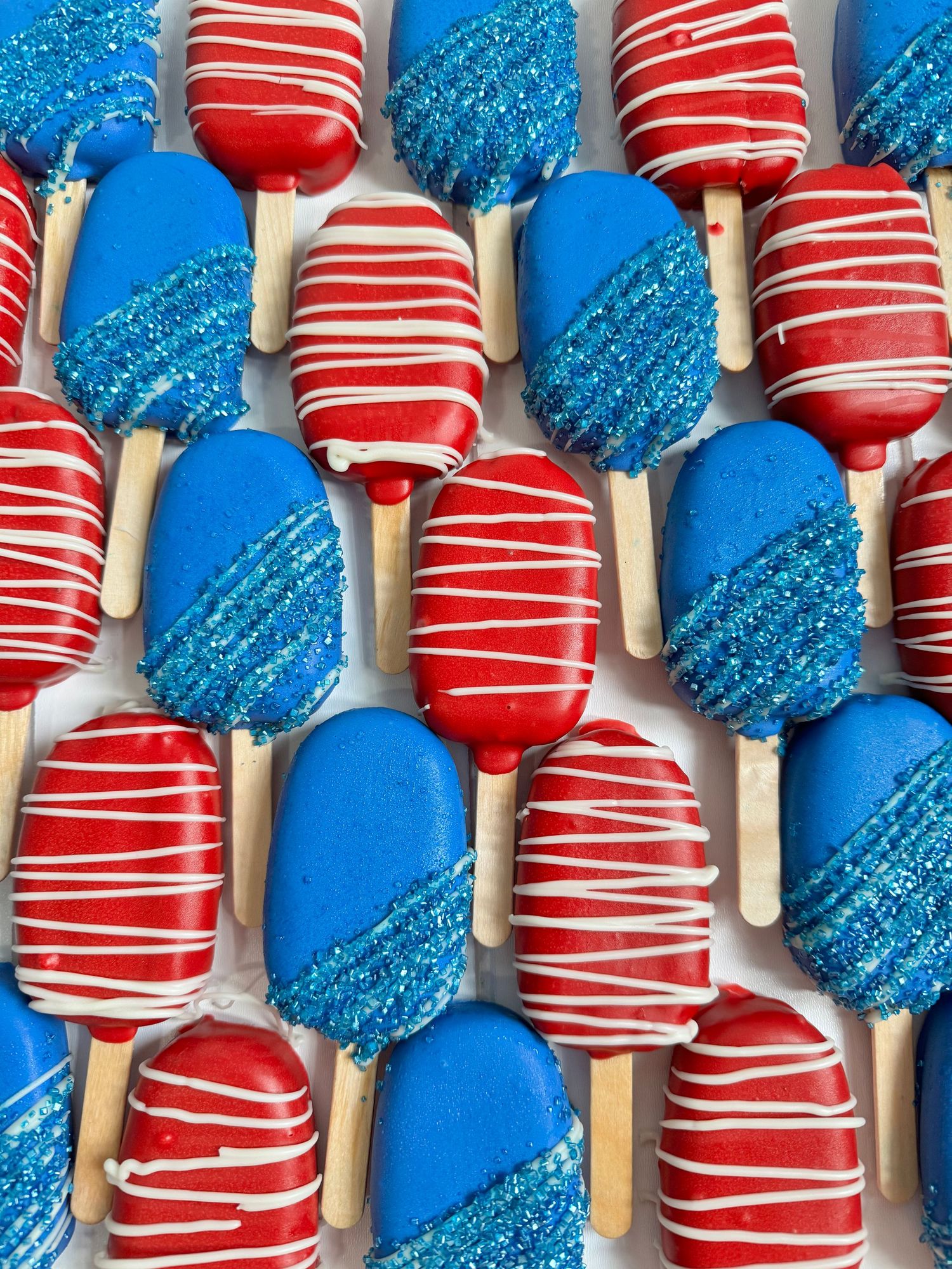 Red and blue cake pops with white drizzle and sparkly sugar coating displayed in rows.
