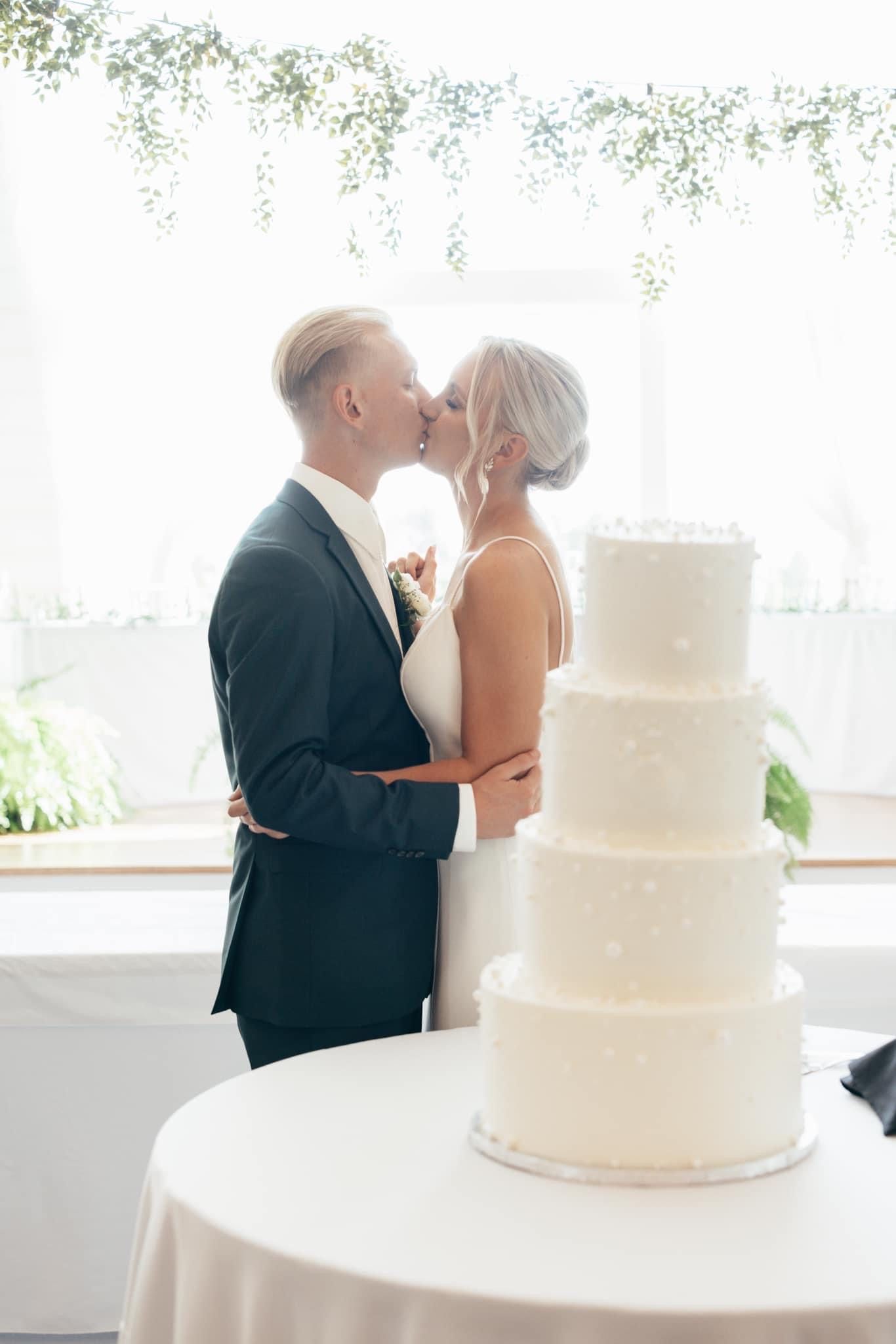 Classic white tiered wedding cake displayed in front of a bright window with greenery.