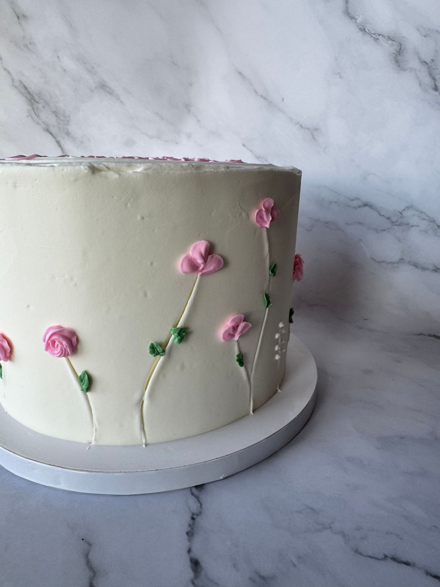 A white frosted cake on a marble surface being decorated with small pink sugar flowers and green leaves.