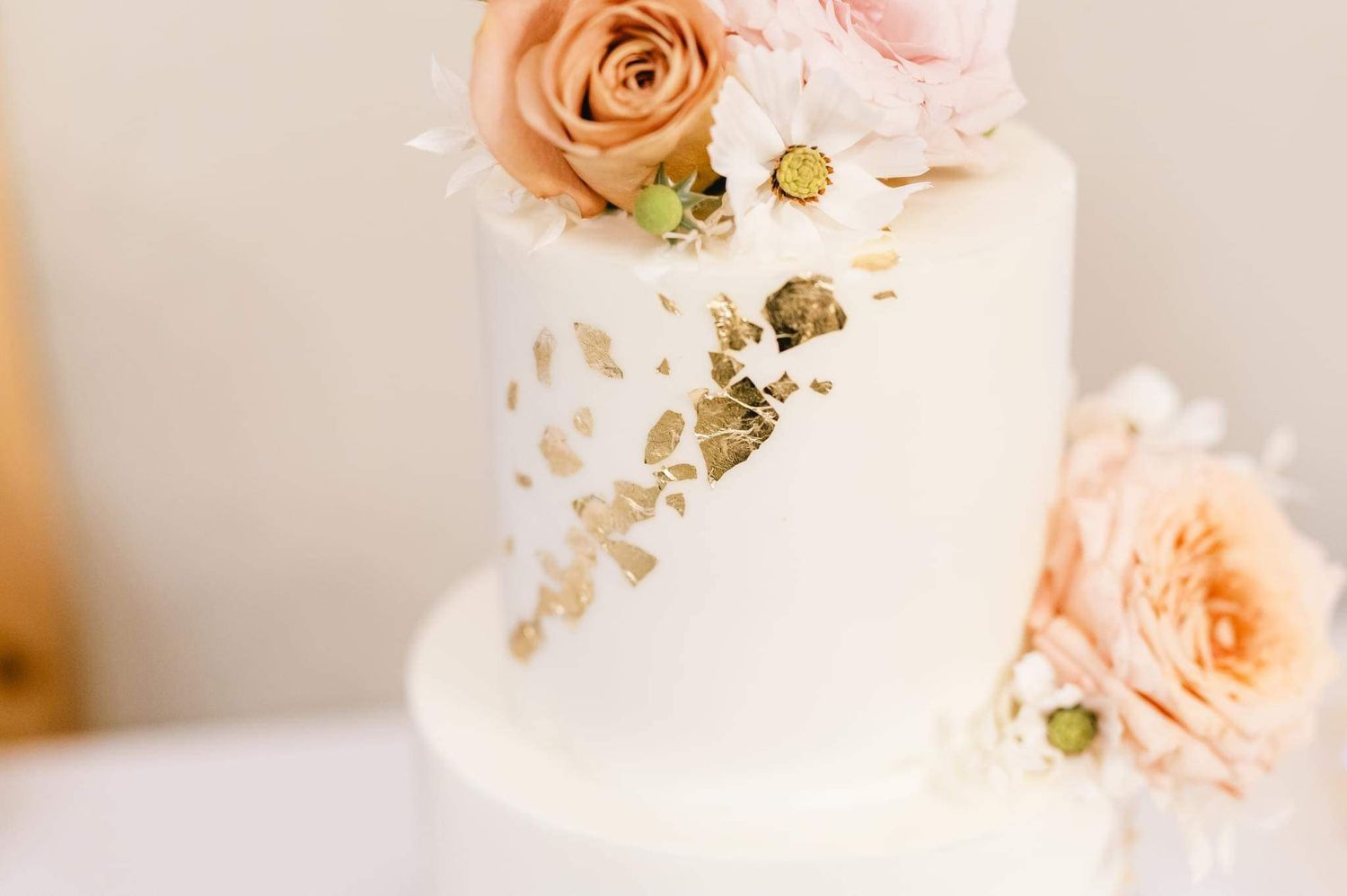 White tiered cake decorated with soft peach roses and white daisies against a neutral background.
