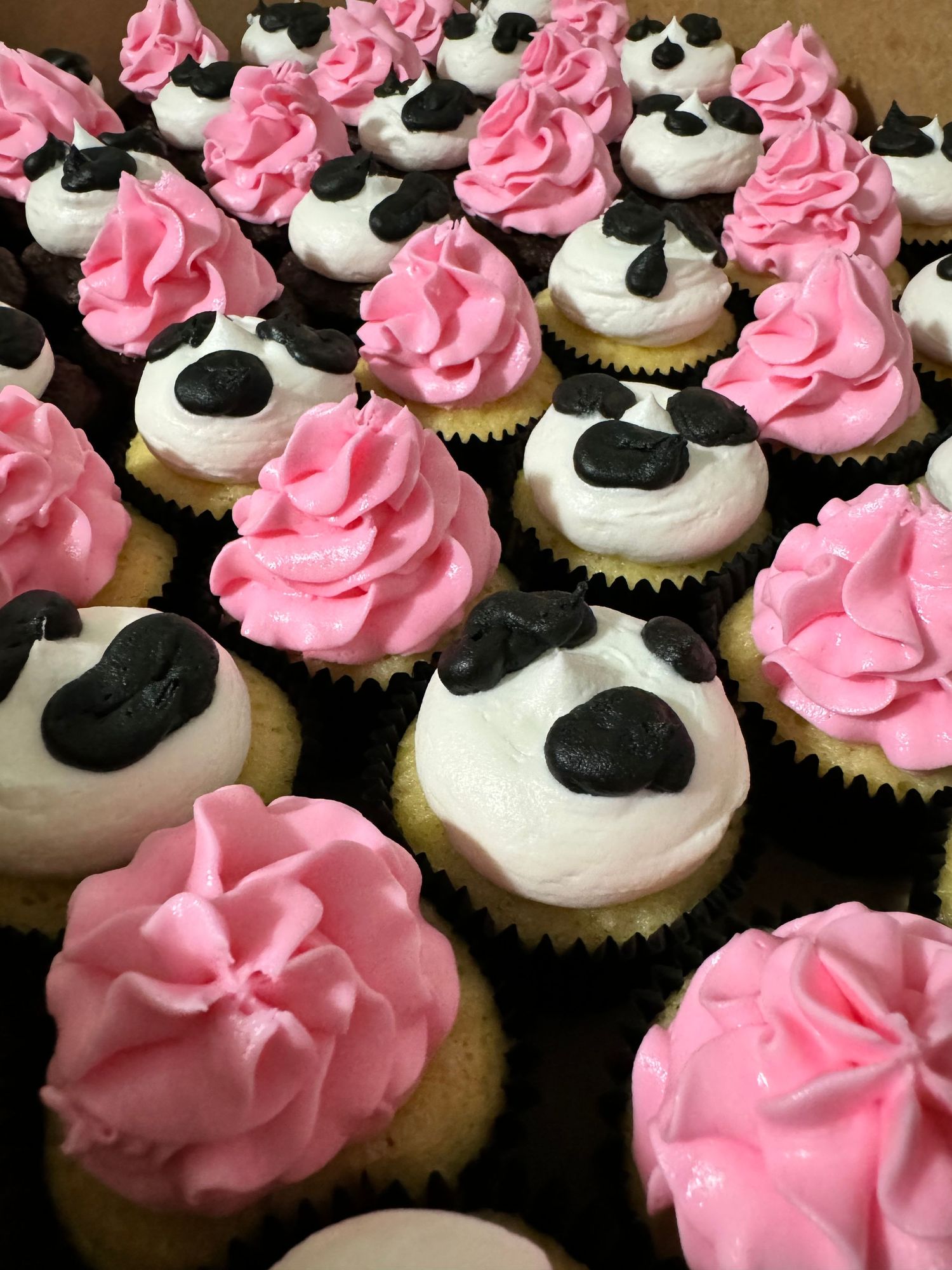Pink frosted cupcakes with black accents arranged in rows on a brown surface.