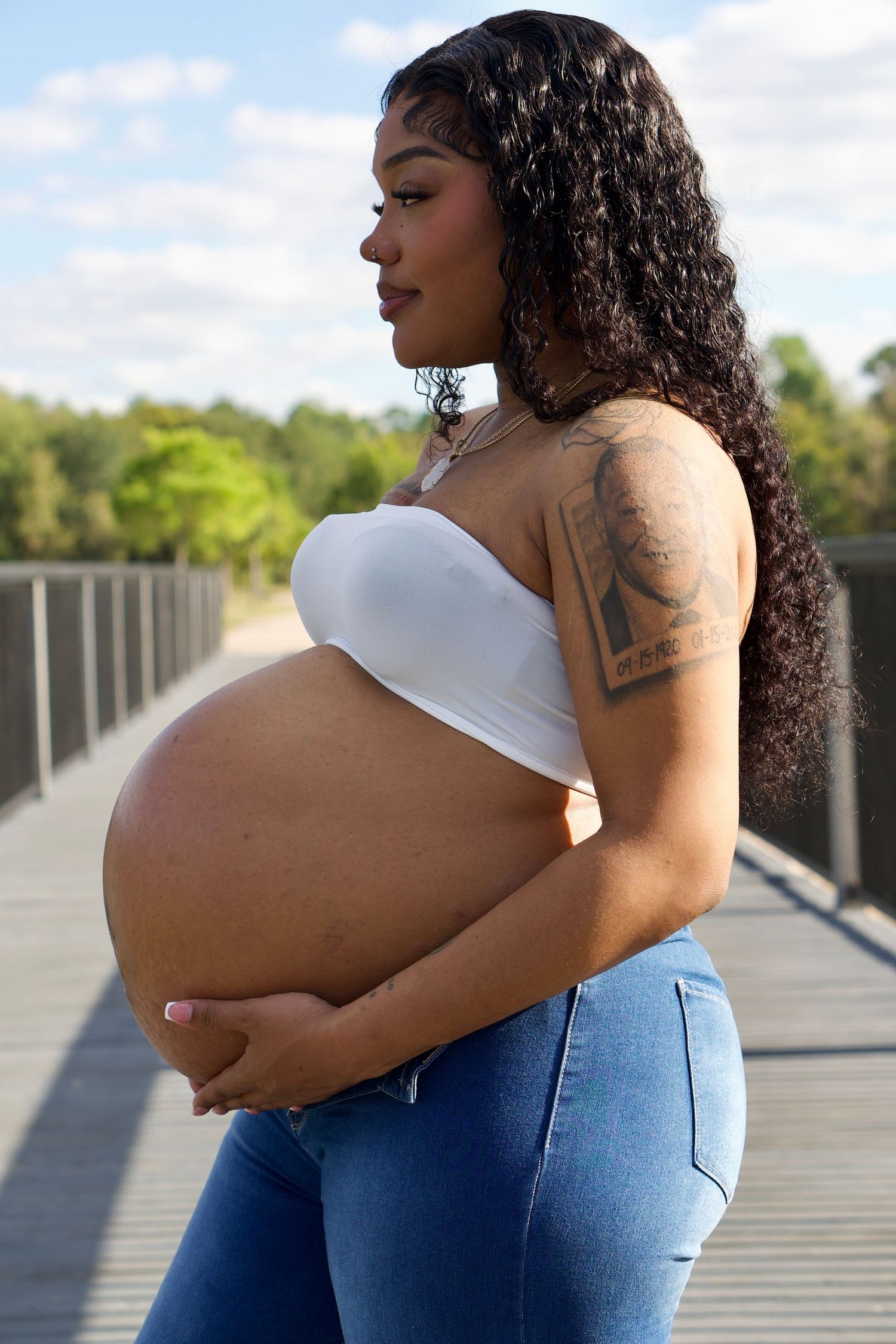 Maternity photoshoot on a wooden boardwalk with blue jeans and white top against natural scenery.