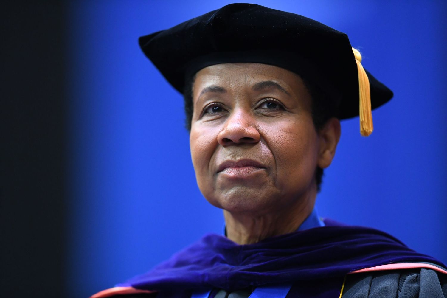 Graduate leader in academic regalia smiling against blue background during commencement.