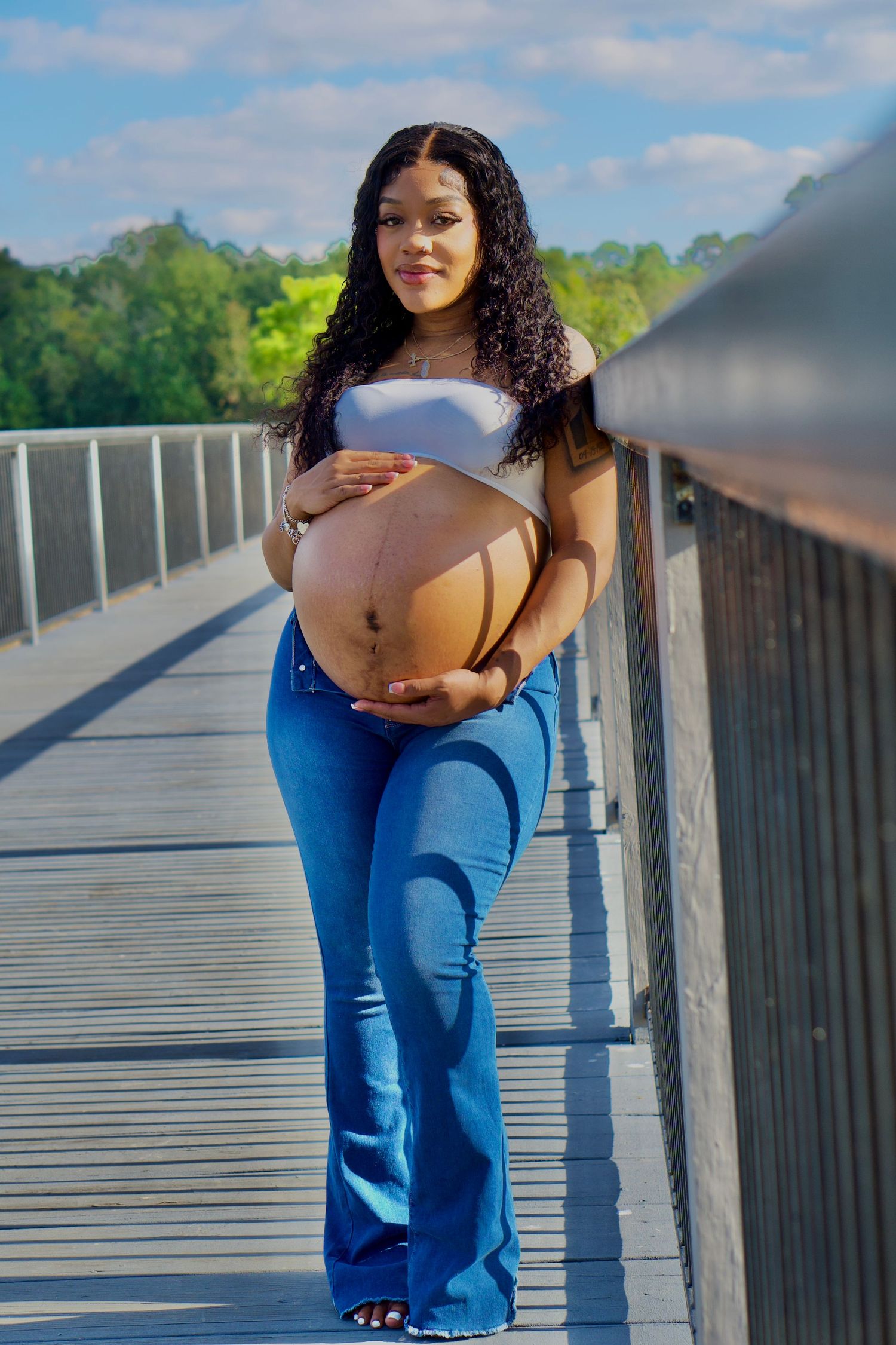 Pregnancy photoshoot captured on wooden walkway with scenic water and trees in background.