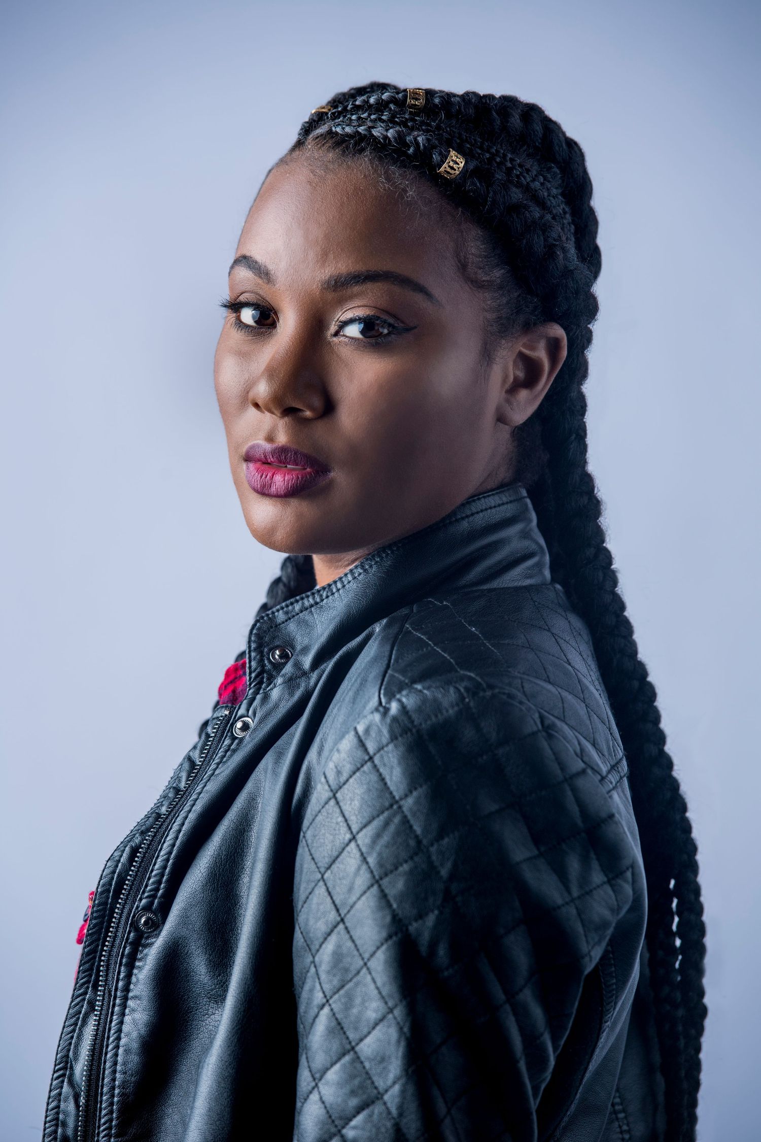 Professional portrait of a model wearing a black leather jacket and braided hairstyle against a blue background.