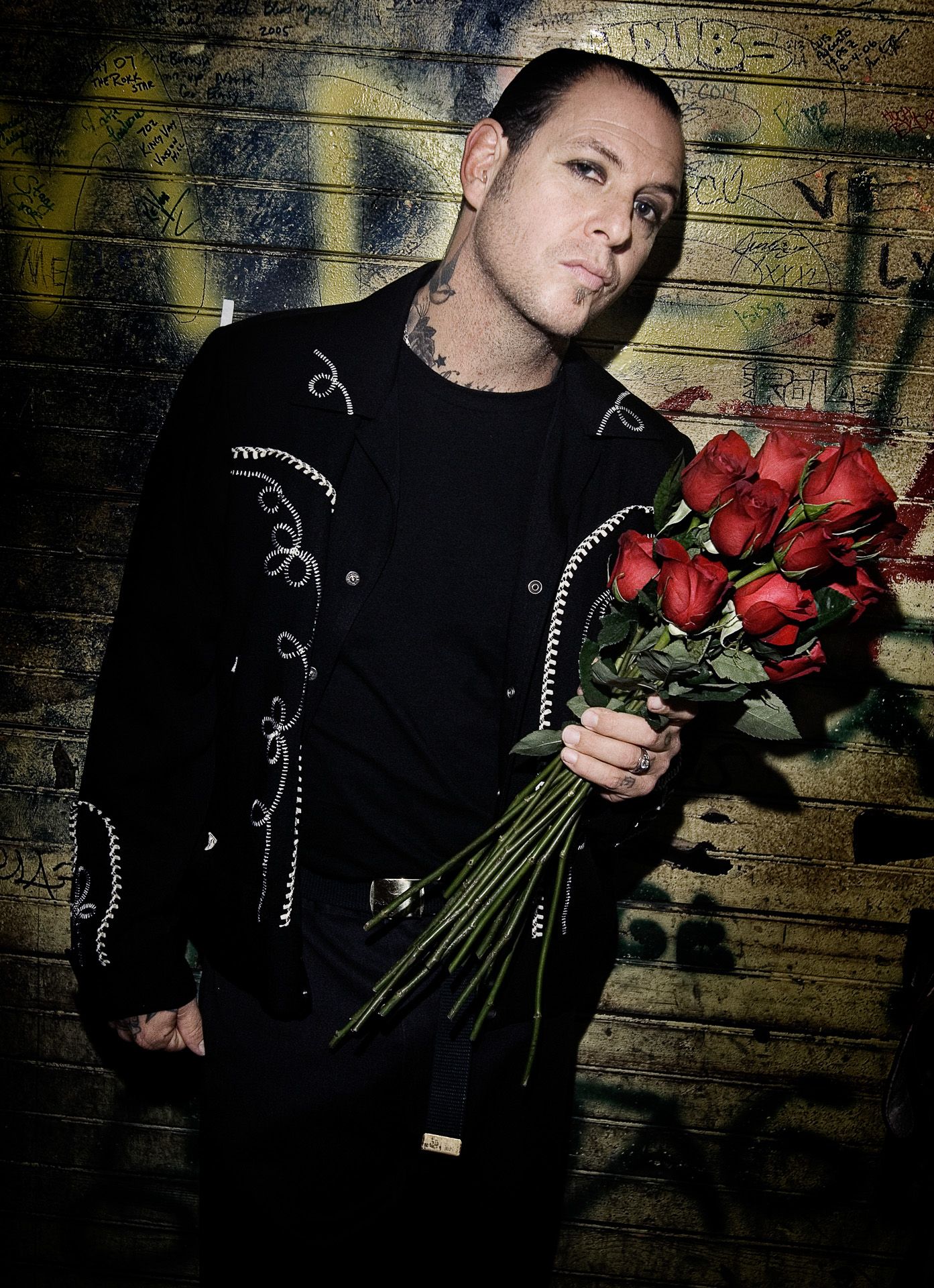 Musician in black embellished jacket holding a bouquet of red roses against weathered wooden wall.