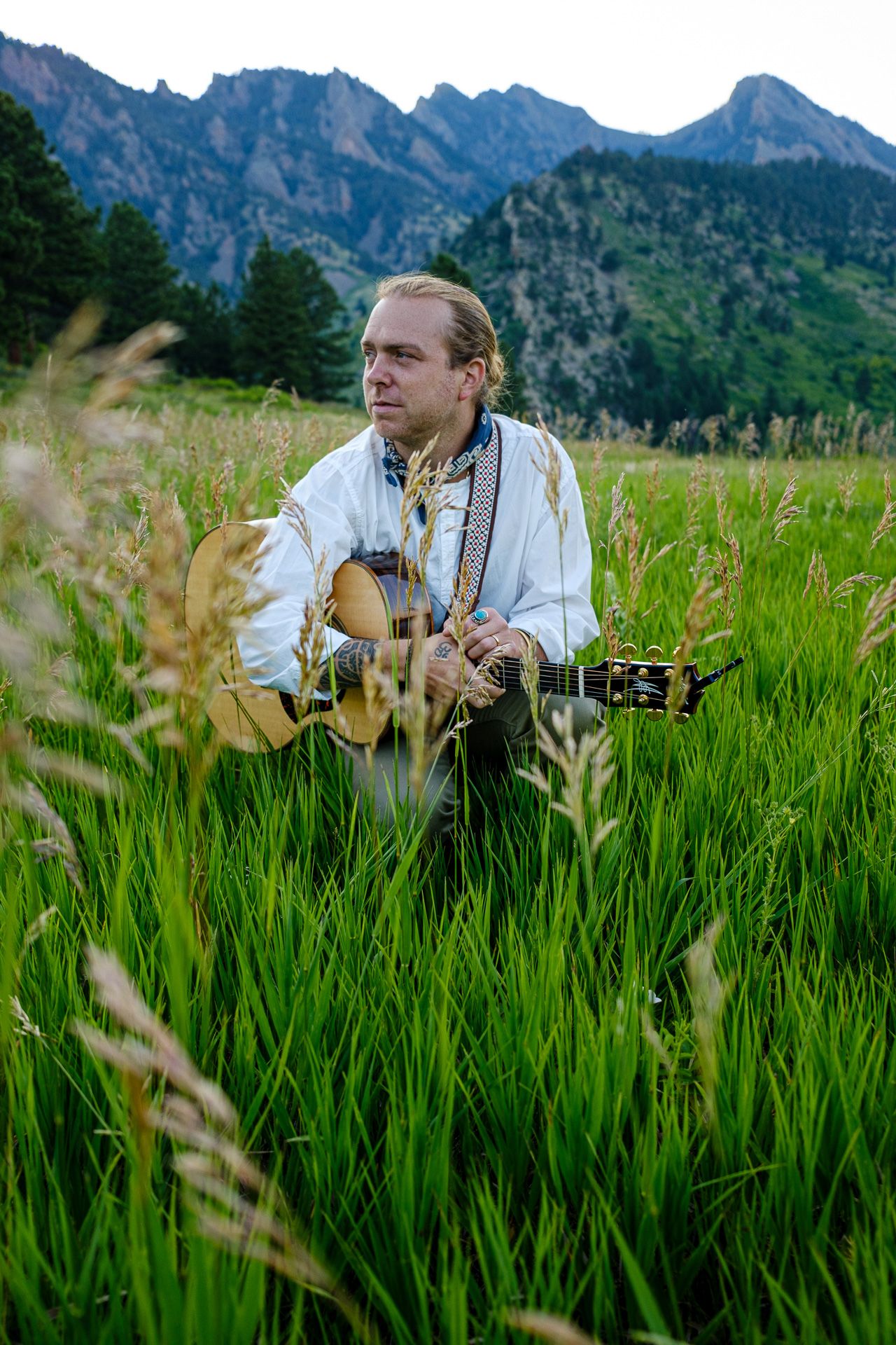 Folk musician plays guitar while sitting in tall grass with mountain range backdrop.