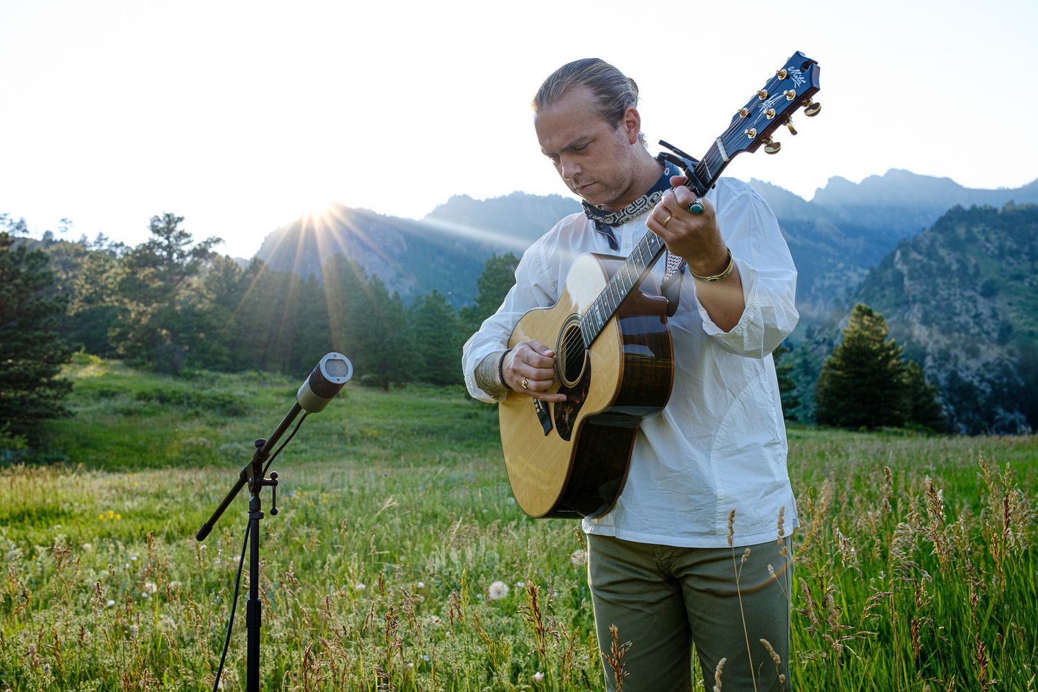 Musician performs with acoustic guitar in mountain meadow during golden hour.