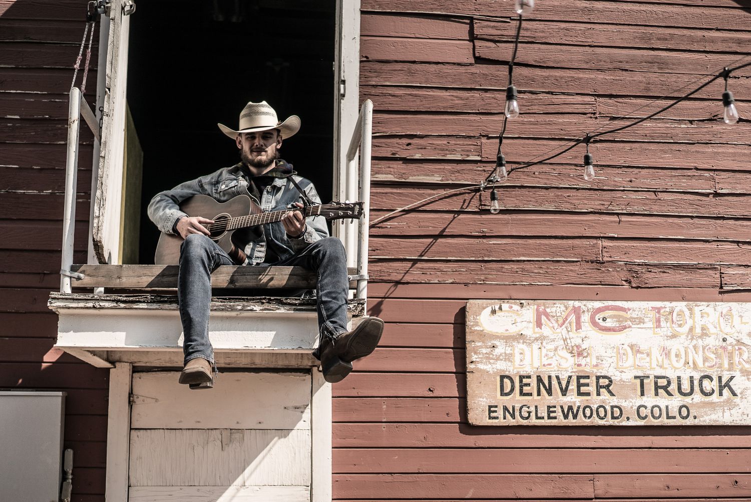 Musician in western wear plays guitar while sitting in window of old Denver Truck building.