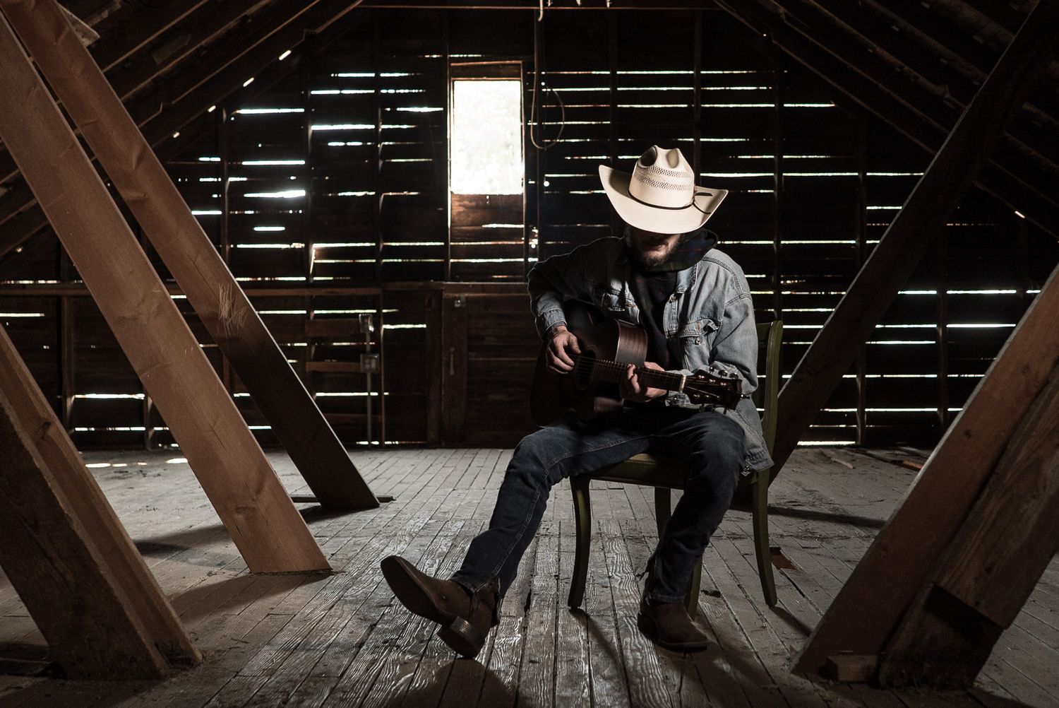 Silhouetted cowboy figure sitting in rustic barn attic with dramatic lighting through wooden slats.