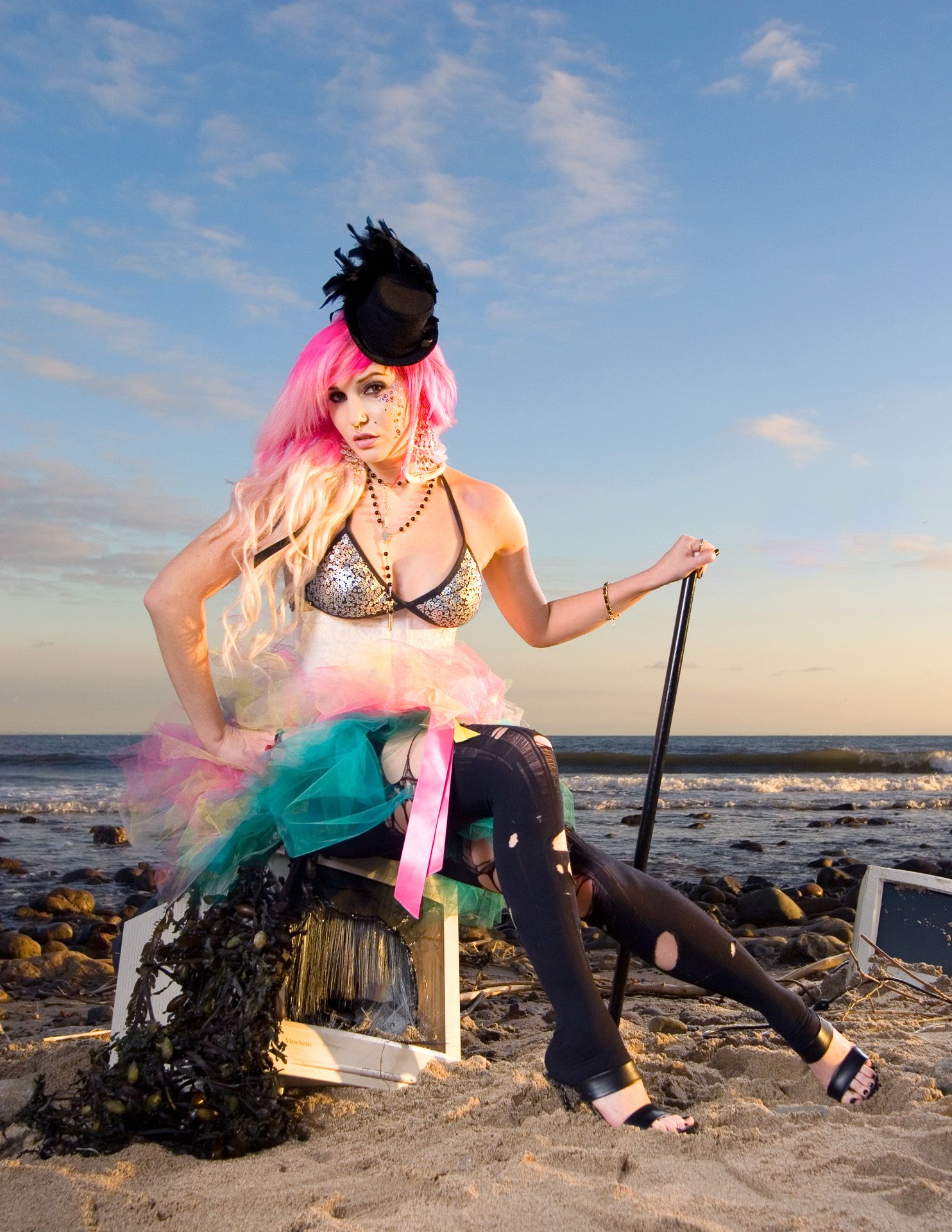 Colorful fantasy-styled figure in steampunk attire poses dramatically on rocky beach at sunset.
