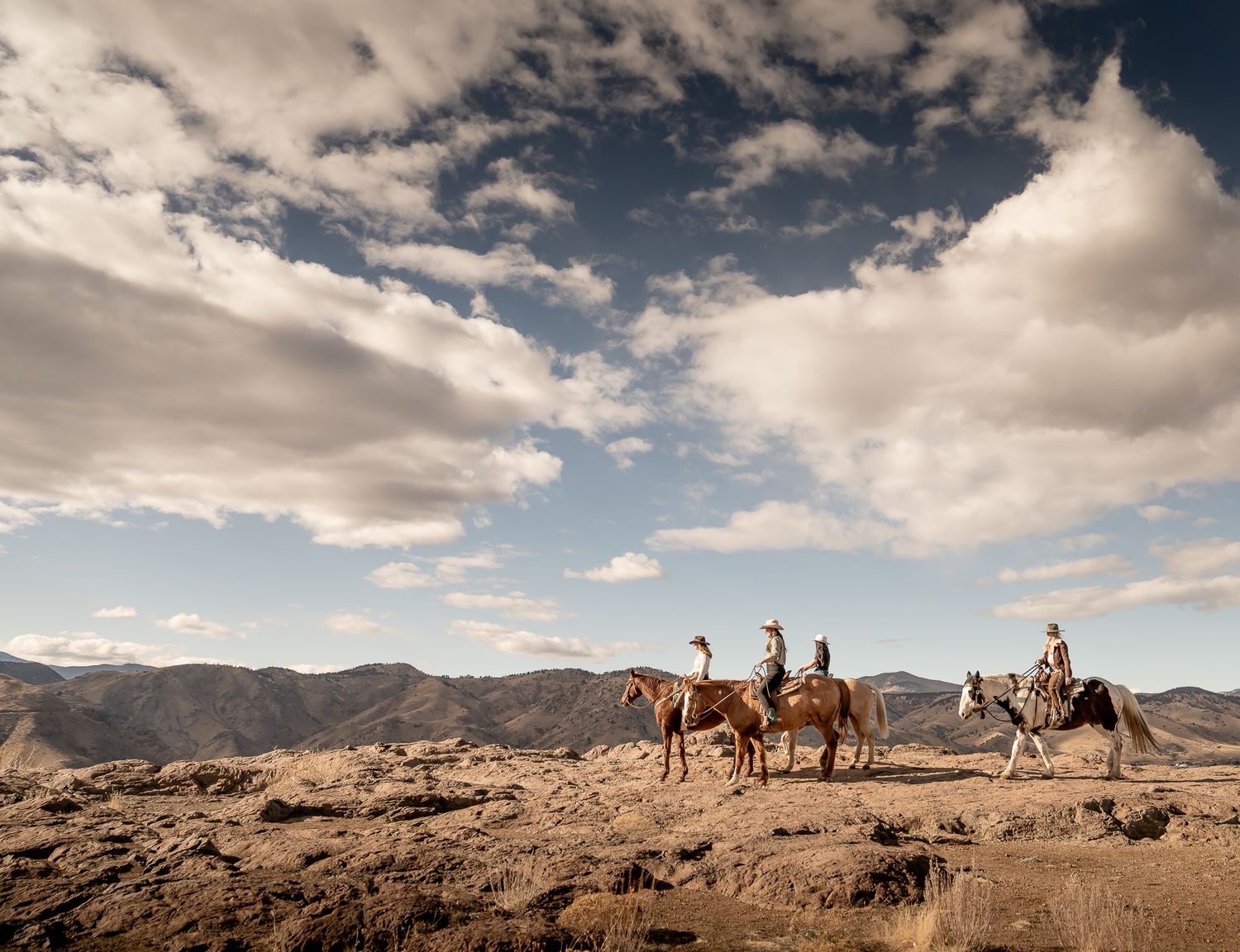 Horseback riders traverse rocky desert terrain under dramatic cloudy sky.