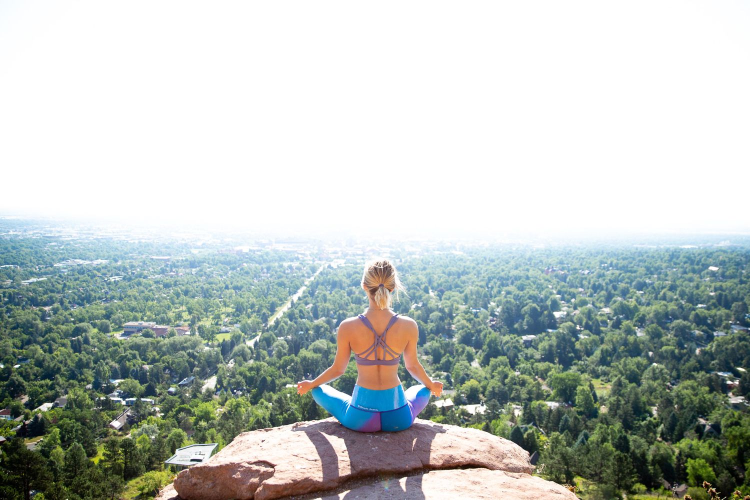 Person meditating on rocky overlook with sprawling city view below.