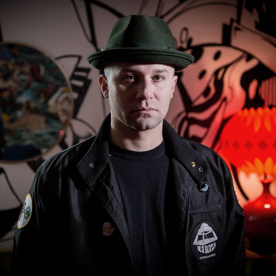 Person in dark clothing and cap photographed dramatically against red backlit urban setting.