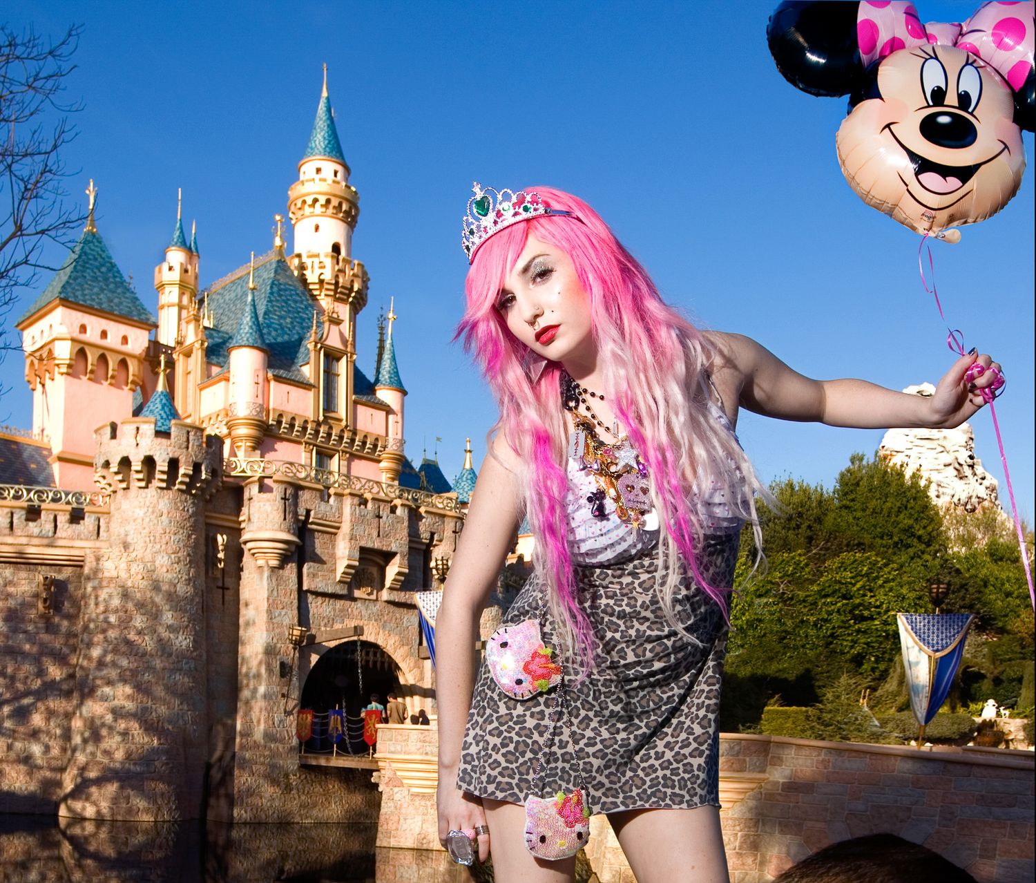 Vibrant pink-haired figure posing playfully in front of Disneyland castle with Minnie Mouse balloon visible in background.