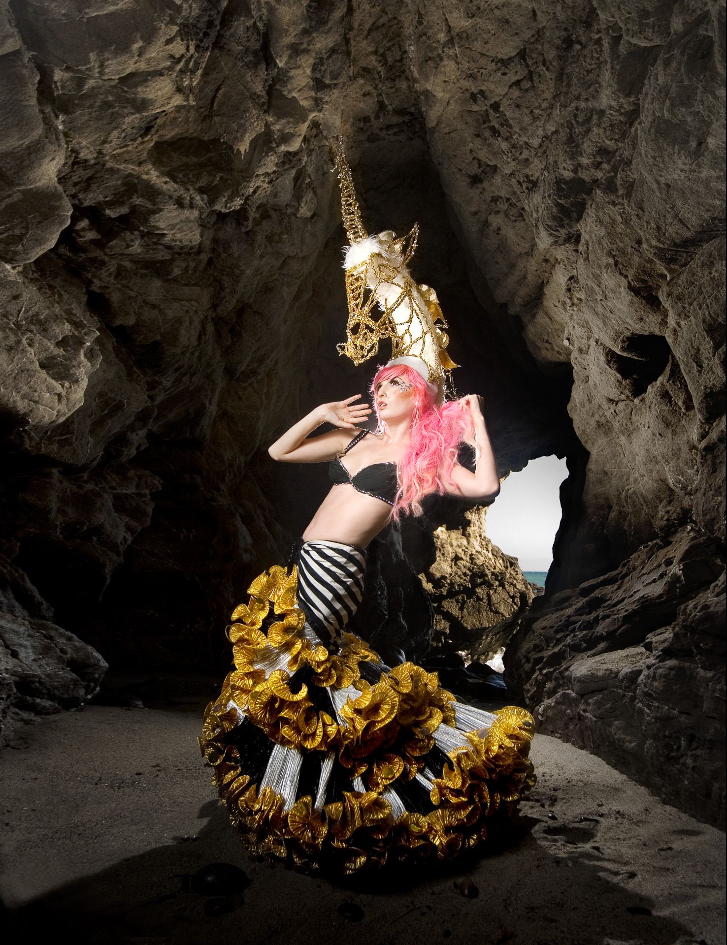 Artistic portrait featuring gold unicorn mask and pink hair against dark cave backdrop.
