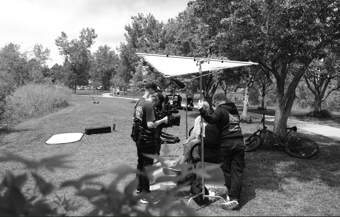 A group of people gather outdoors at a picnic area with trees and grassy spaces visible in the background.