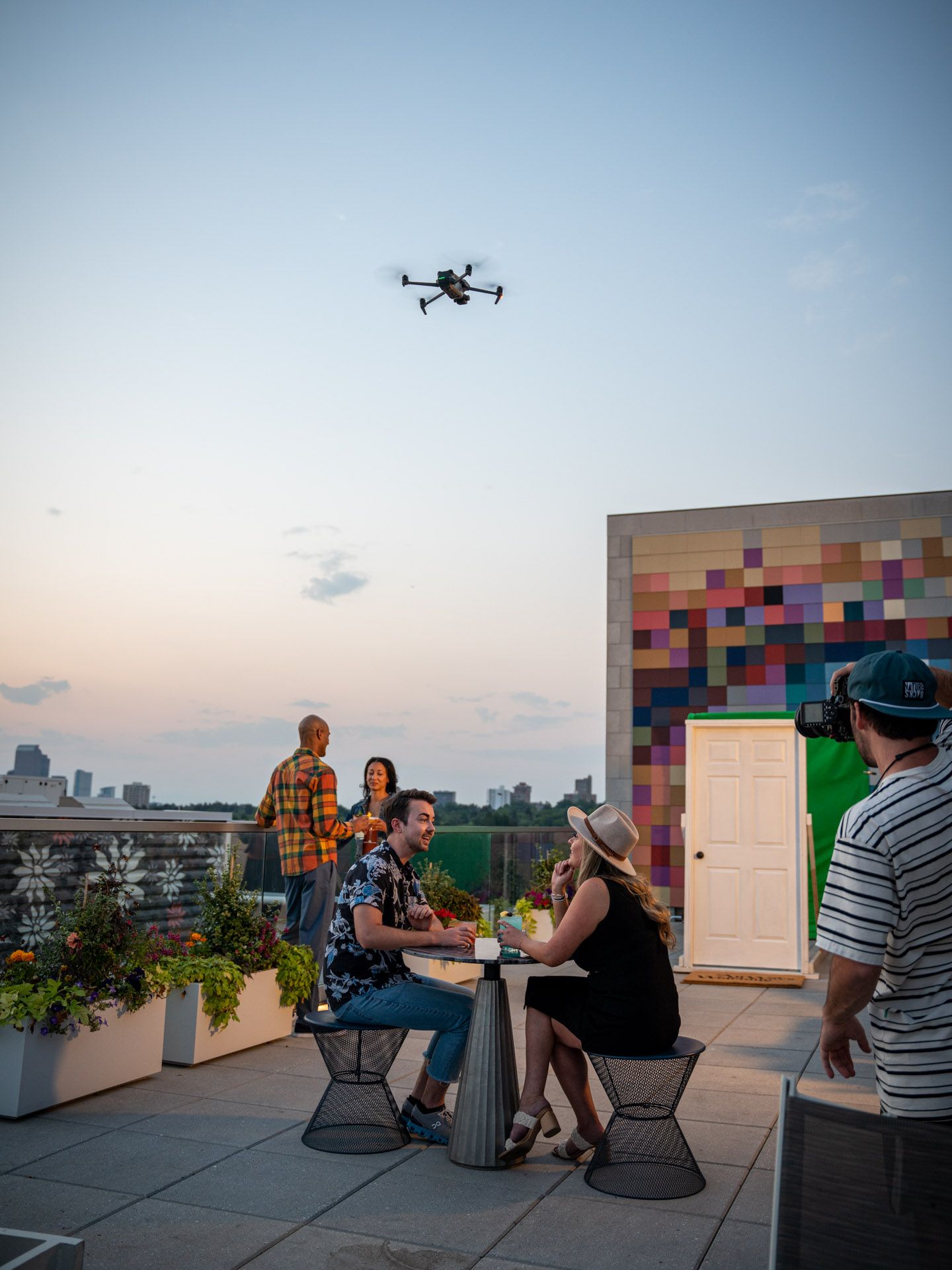 Film crew and equipment set up on a rooftop with city skyline and drone in background at dusk.
