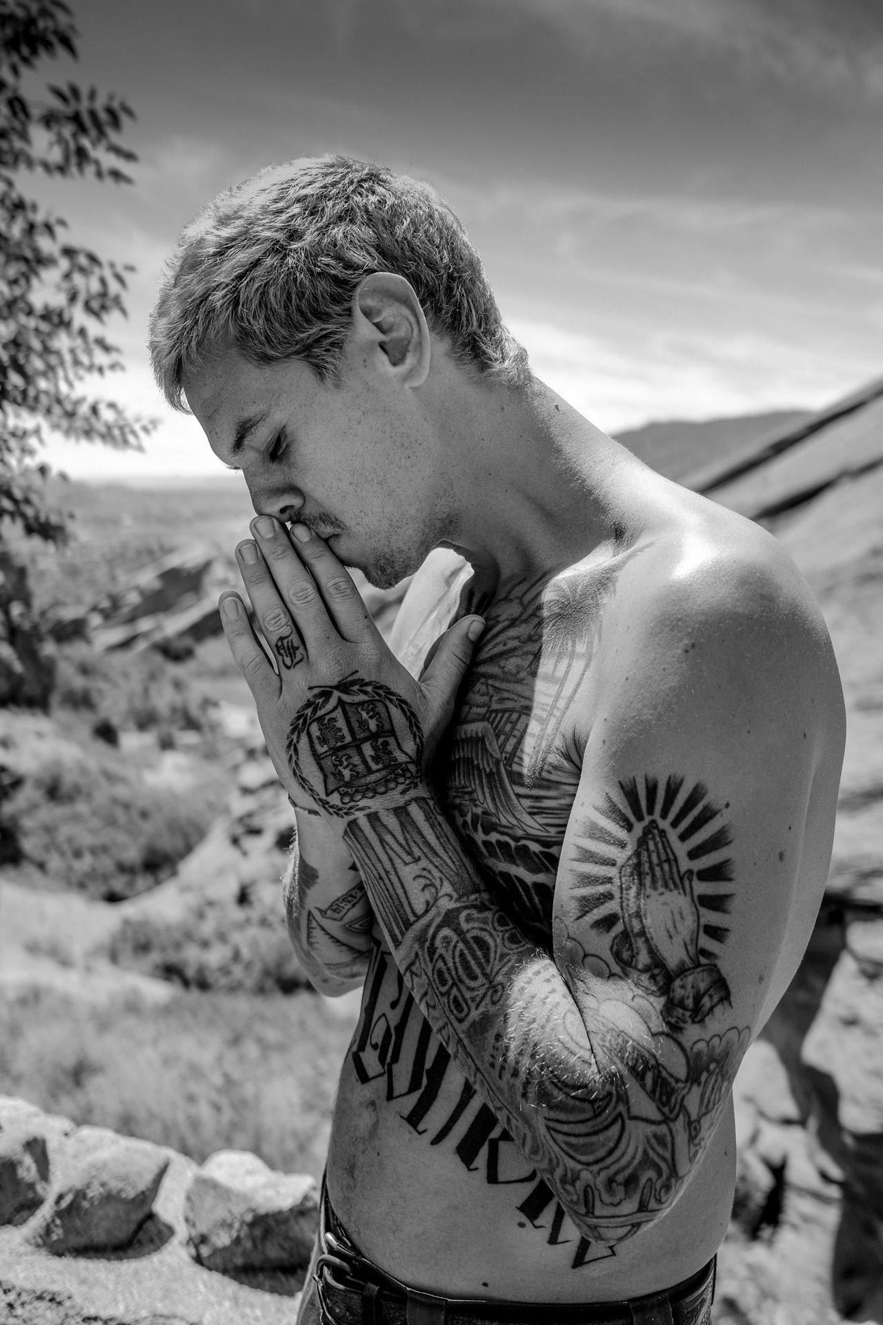 Black and white portrait of tattooed figure in prayer pose against mountainous backdrop.