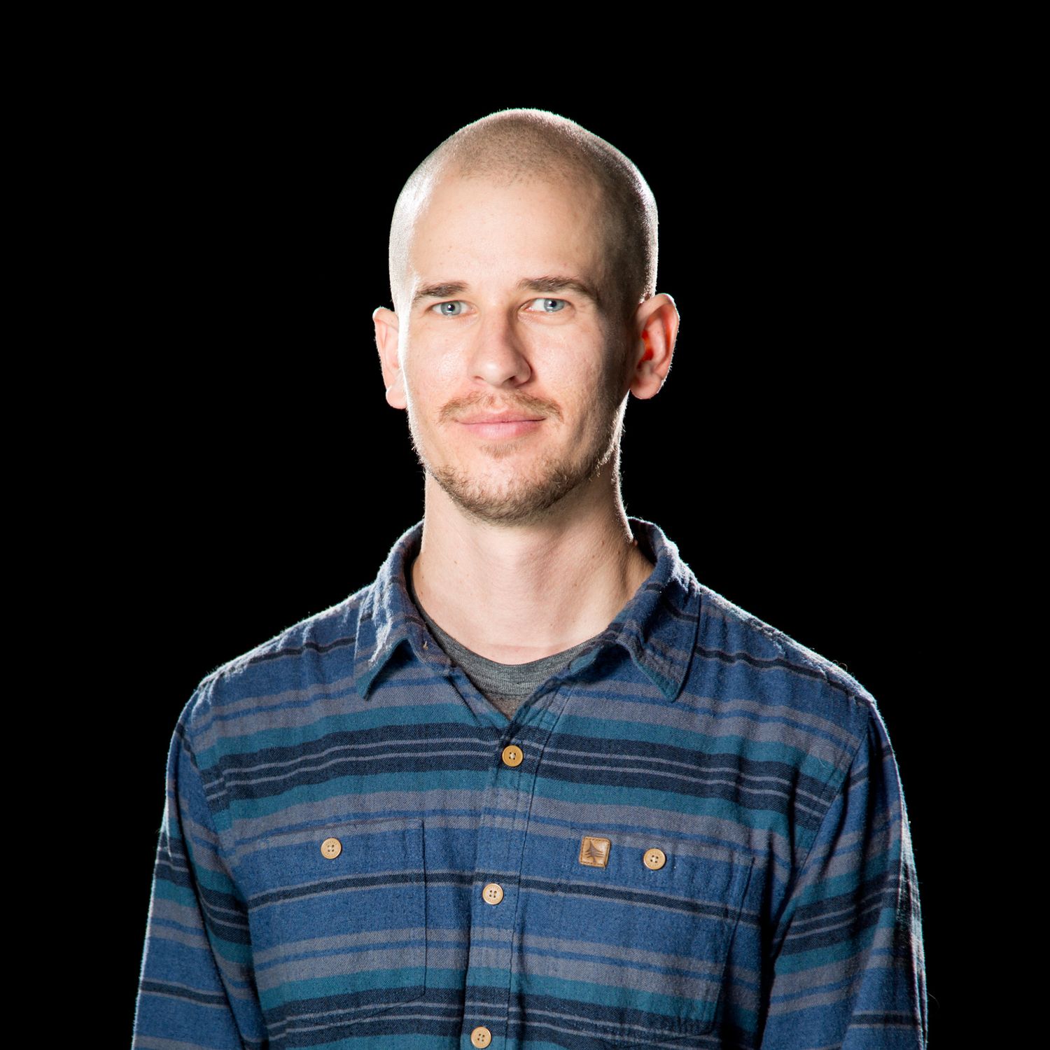 A person wearing a blue and grey plaid button-up shirt stands against a black background.
