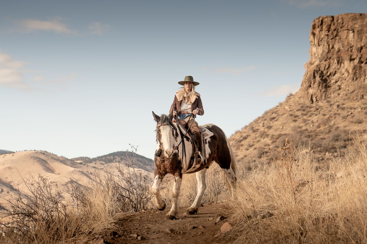 A figure on horseback rides along a desert trail with rugged cliffs and mountains in the backdrop at sunset.