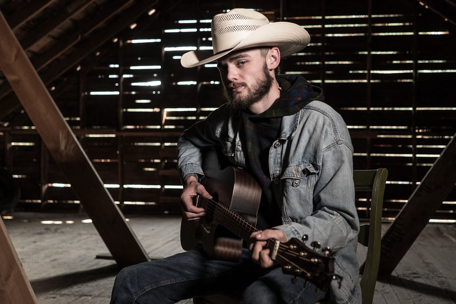Country musician in cowboy hat plays acoustic guitar in rustic barn setting.