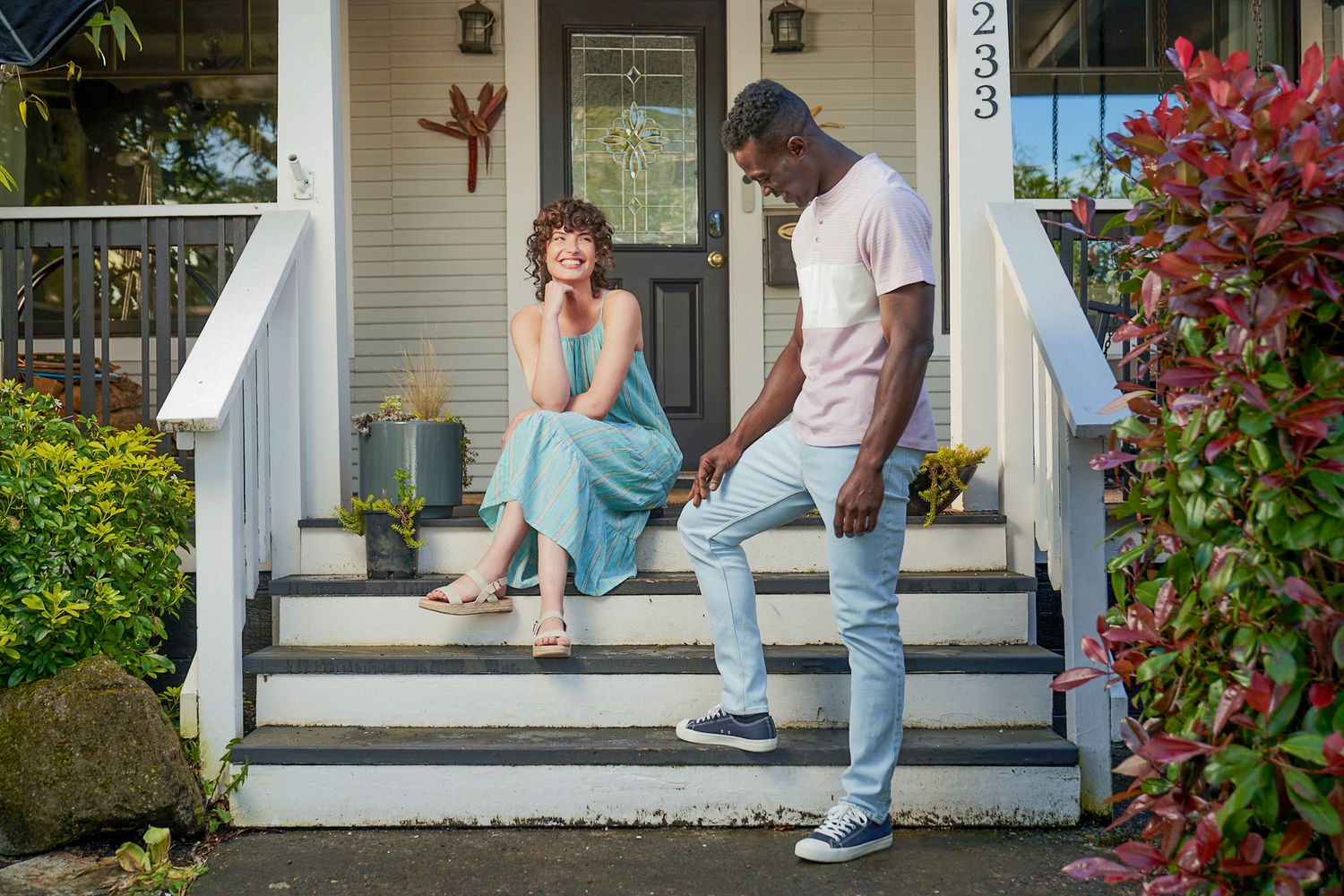 Two friends chatting on white porch steps of traditional home with flowering bushes.