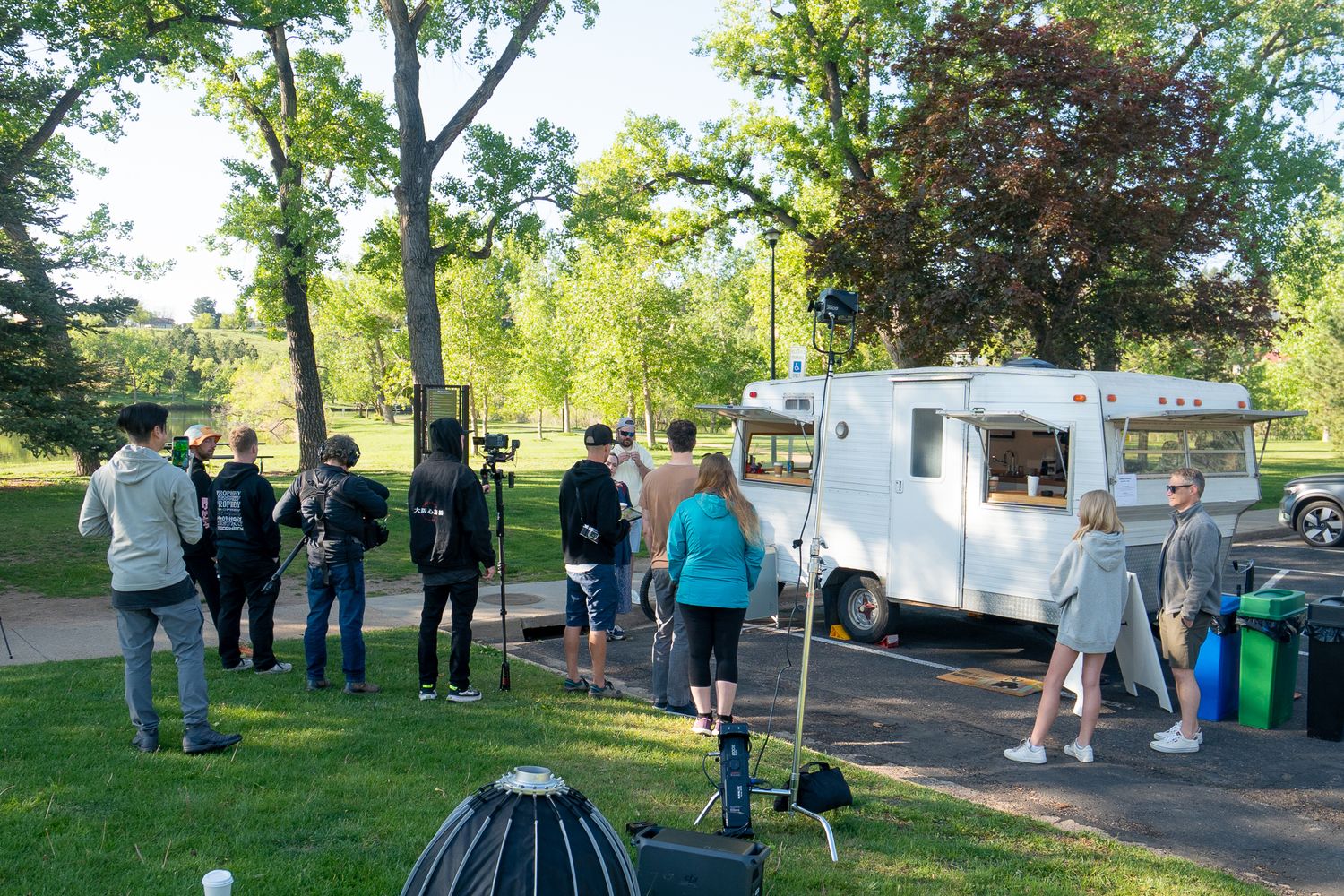 People gather outdoors in a park setting with white tents and equipment visible during a daytime event.