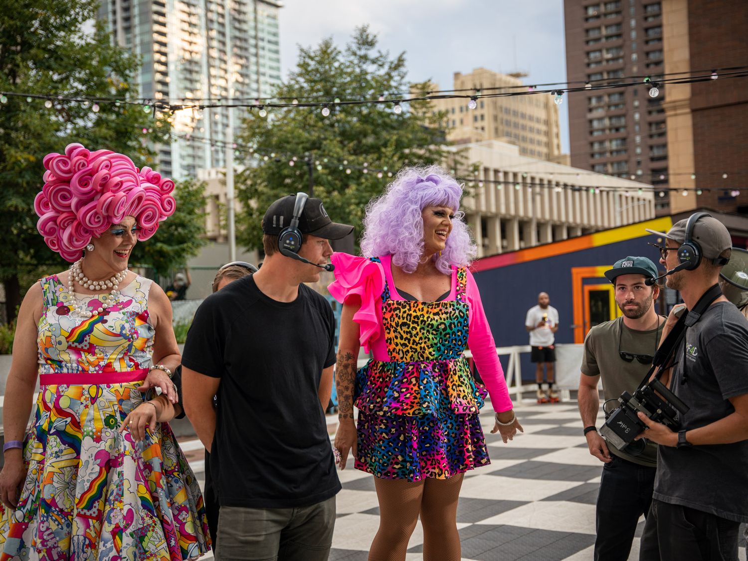 Performers in vibrant drag costumes posing with film crew on a checkered stage area.