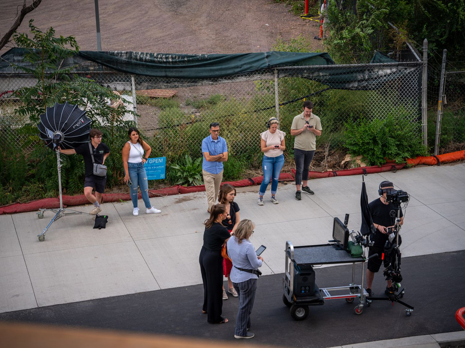 Production crew and equipment set up on outdoor location with crowd in background.
