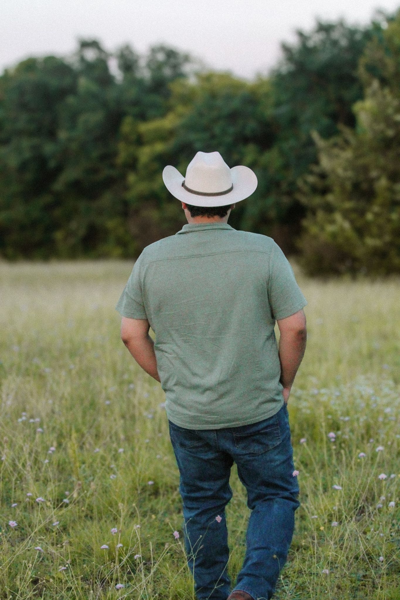 Person wearing cowboy hat and jeans stands in tall grass field during sunset in summer.