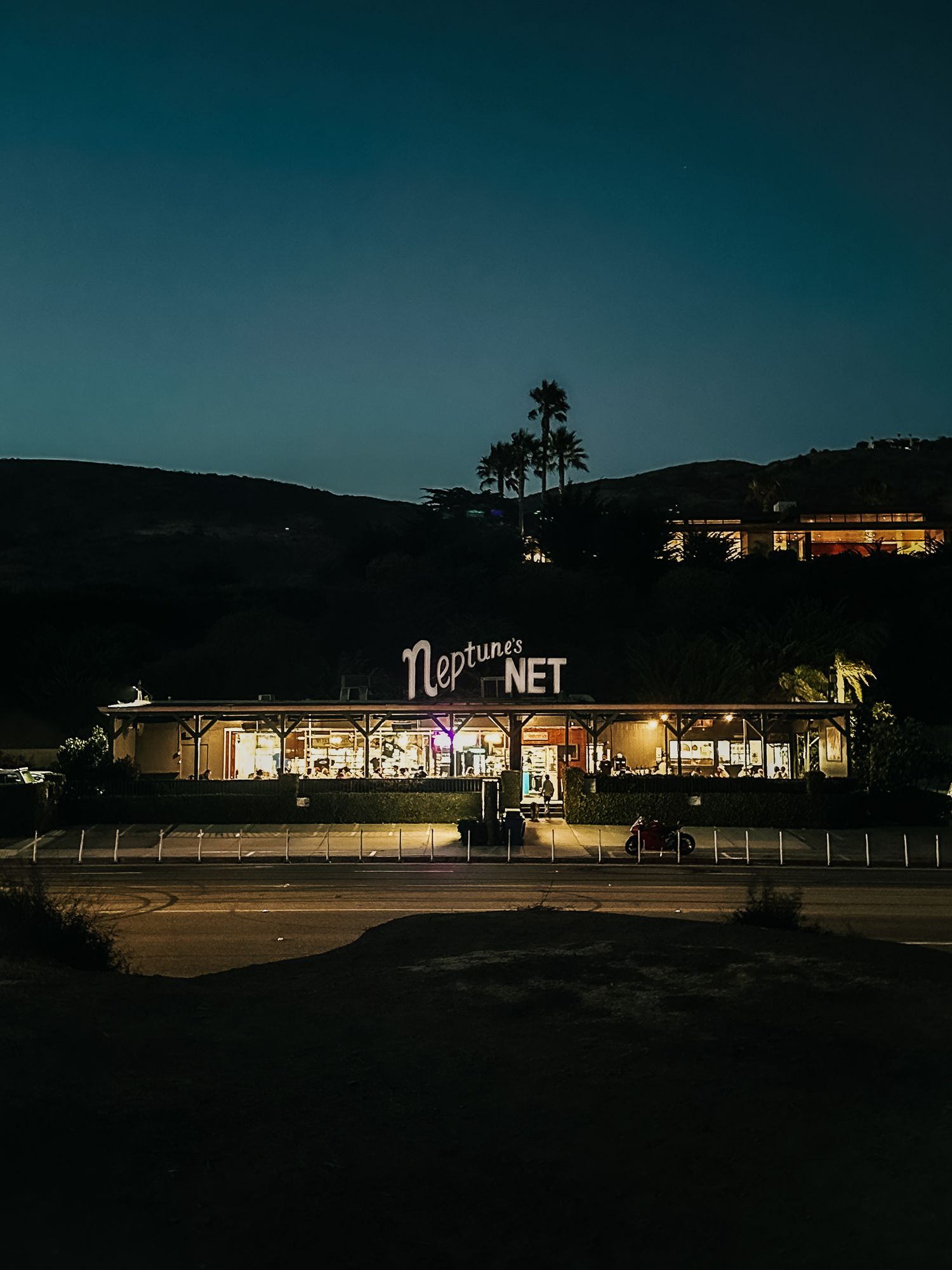 A neon sign for Neptune's Net restaurant glows in the dark against a silhouetted mountain backdrop at dusk.