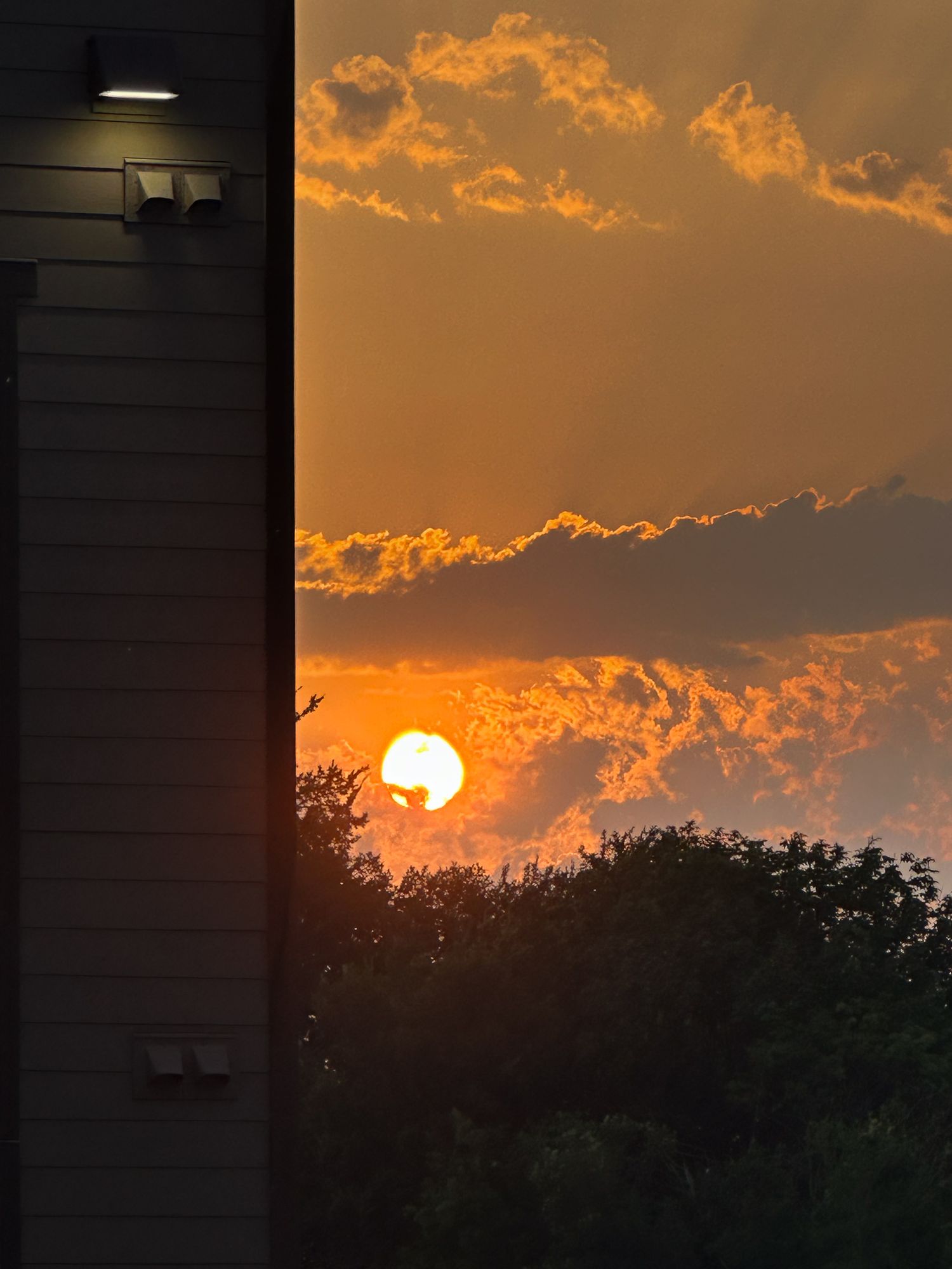 Dramatic orange sunset behind silhouetted trees and buildings with glowing clouds in the twilight sky.