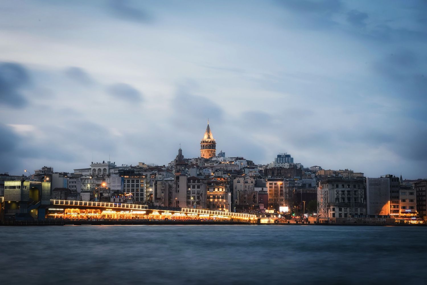 The historic Galata Tower rising above the Istanbul, Turkey skyline, viewed across the illuminated waterfront at twilight.