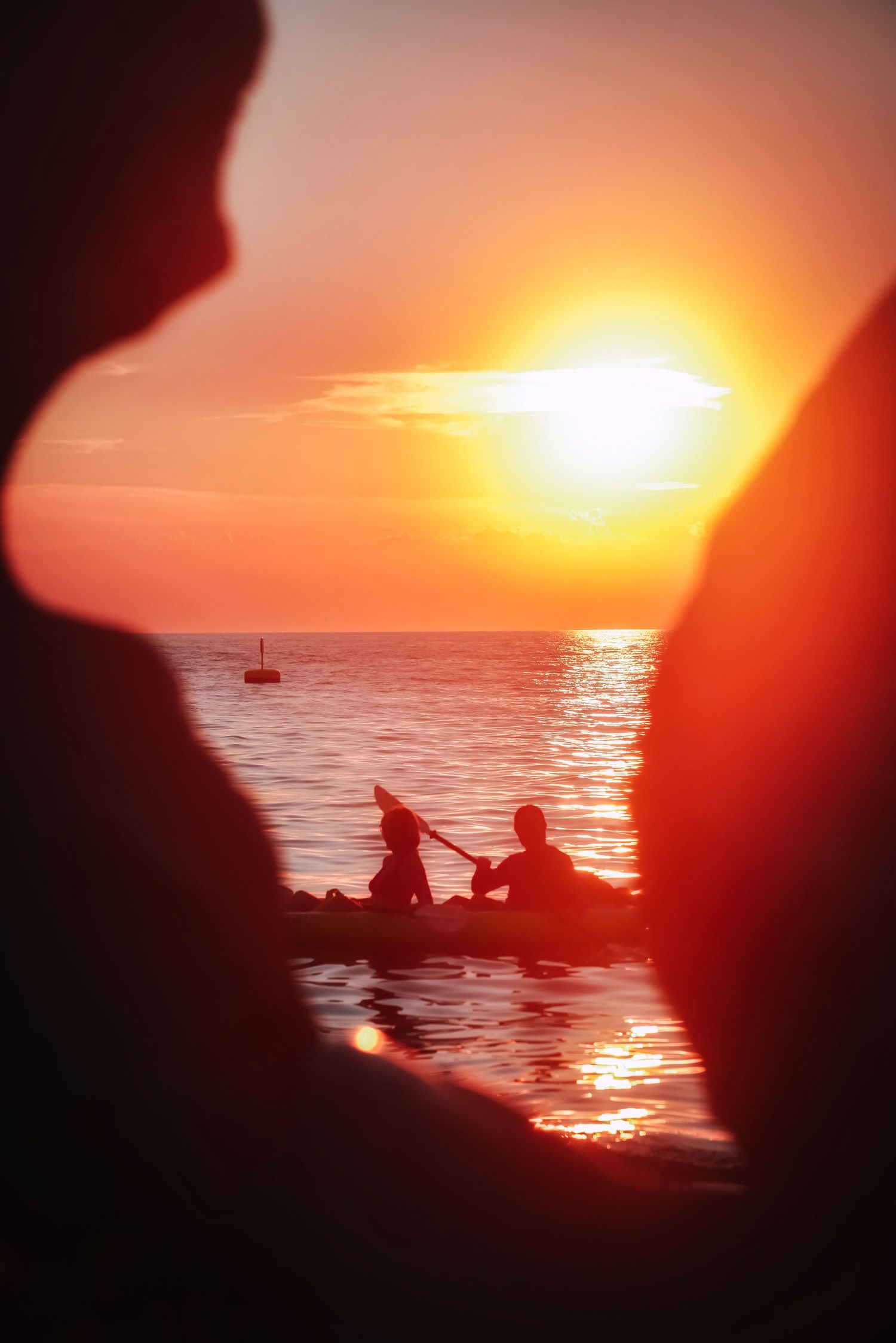 Silhouettes of people kayaking at sunset on the water in Piran, Slovenia, framed by dark foreground silhouettes