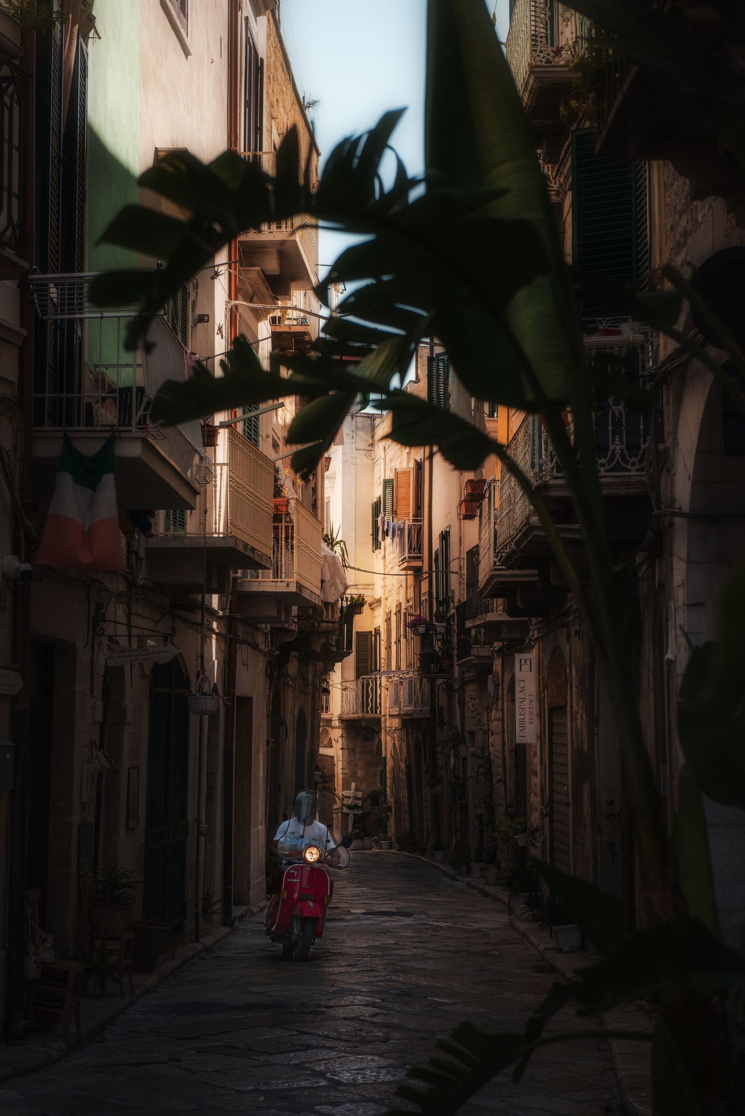 Person riding a red scooter through a narrow, shadowed historic street in Trani, Apulia, Italy.