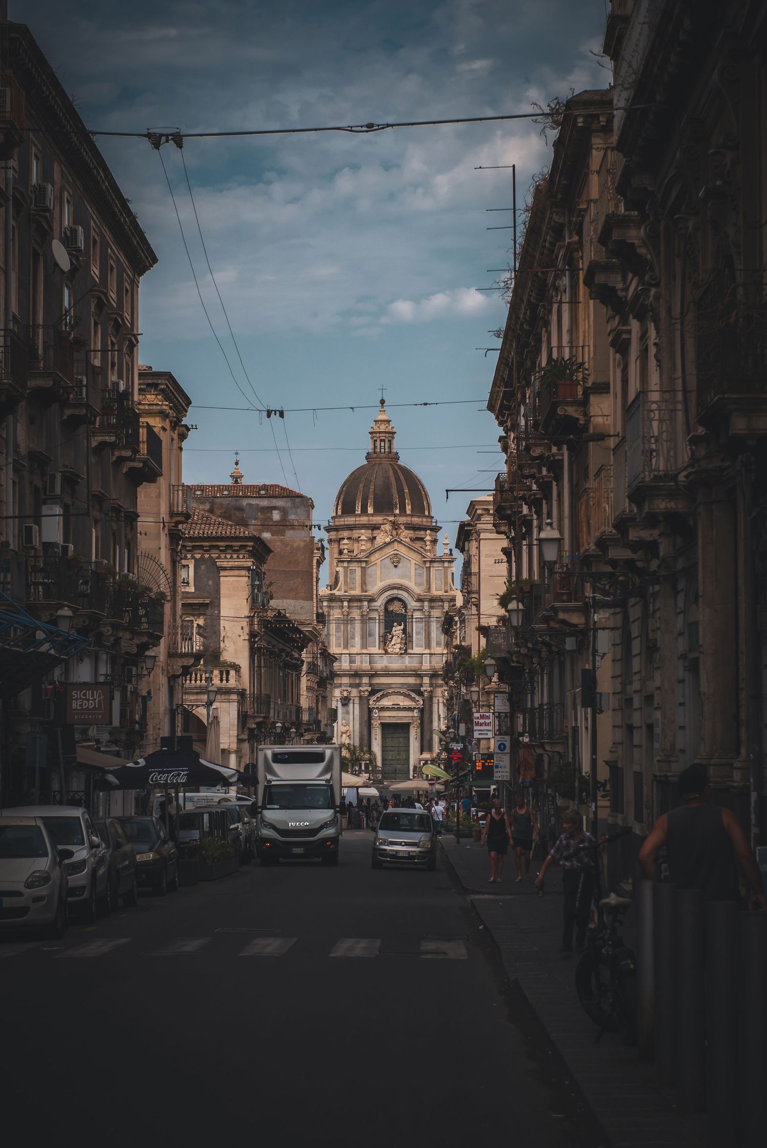 Historic street in Catania, Sicily, Italy, framing a distant view of a baroque domed church and city traffic.