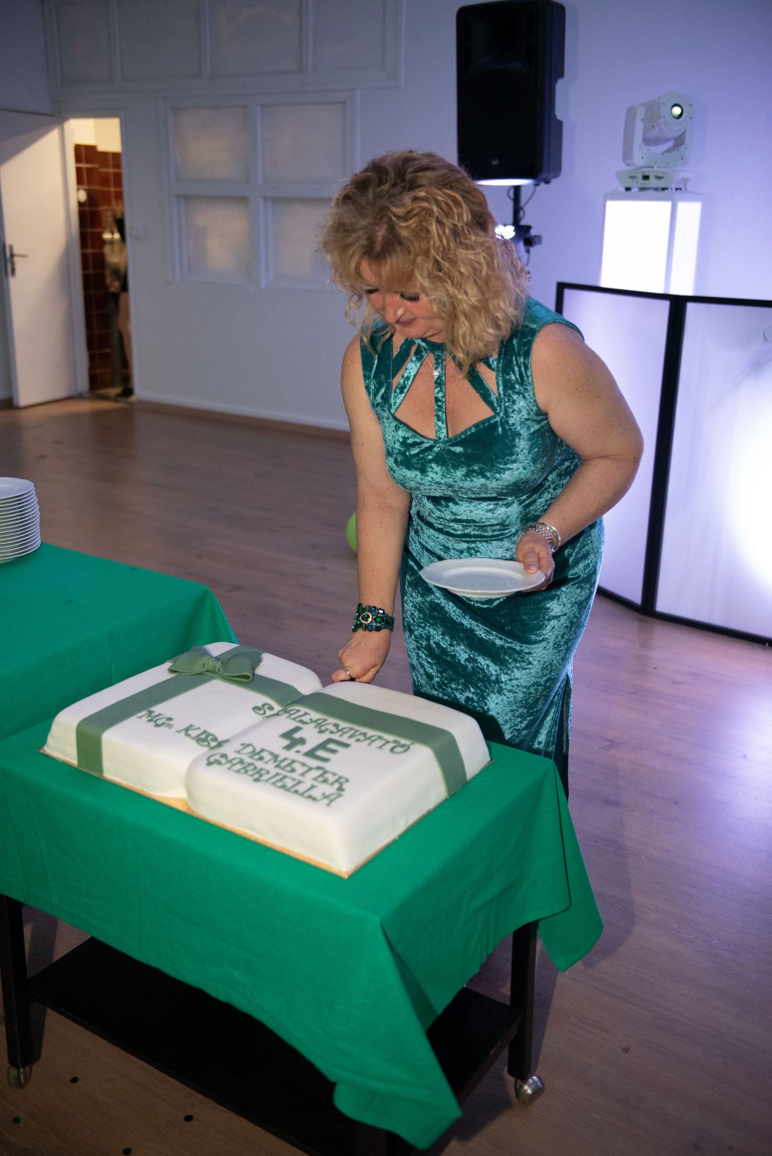 A woman in an elegant green dress cuts a large, book-shaped class cake at a high school prom event.