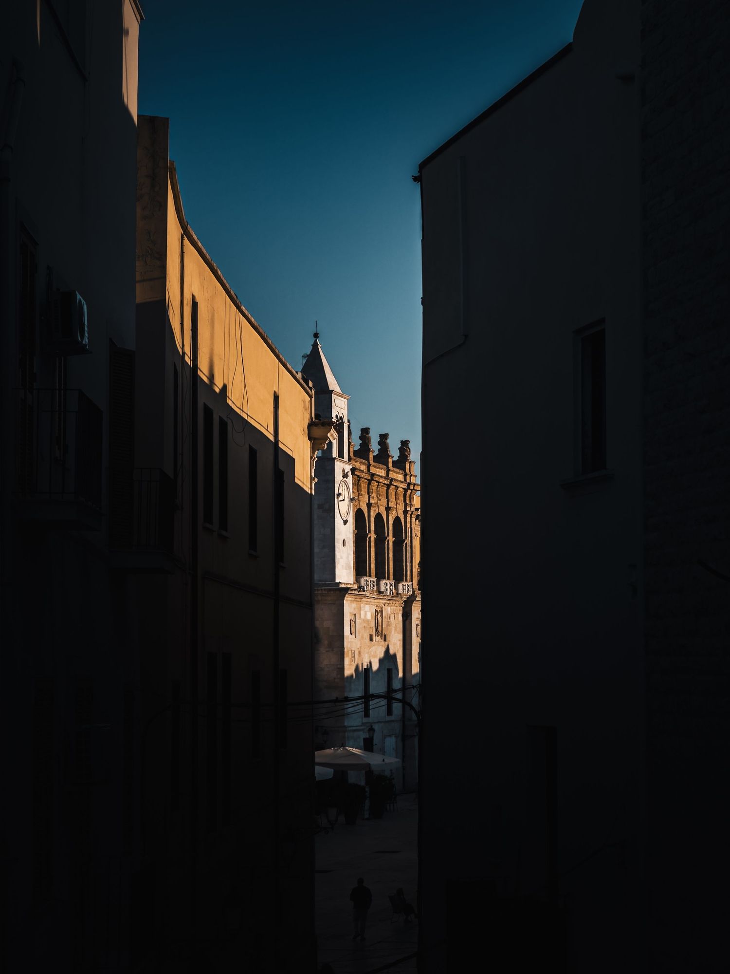 Narrow shaded alleyway in Bari, Apulia, Italy, looking toward a sunlit historic stone bell tower.