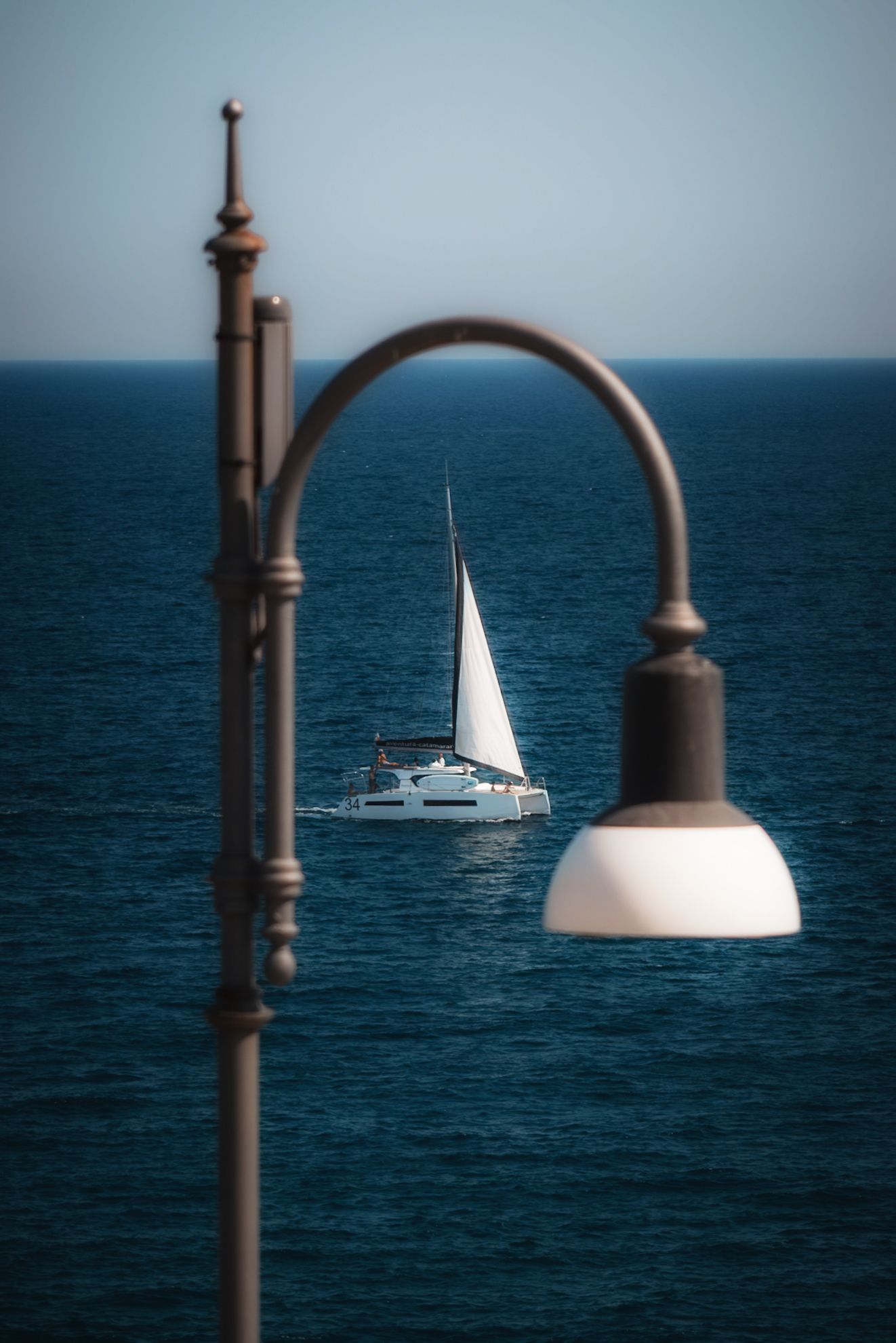 Sailboat on the sea framed by a street lamp, viewed from near the Domenico Modugno statue in Polignano a Mare, Italy.