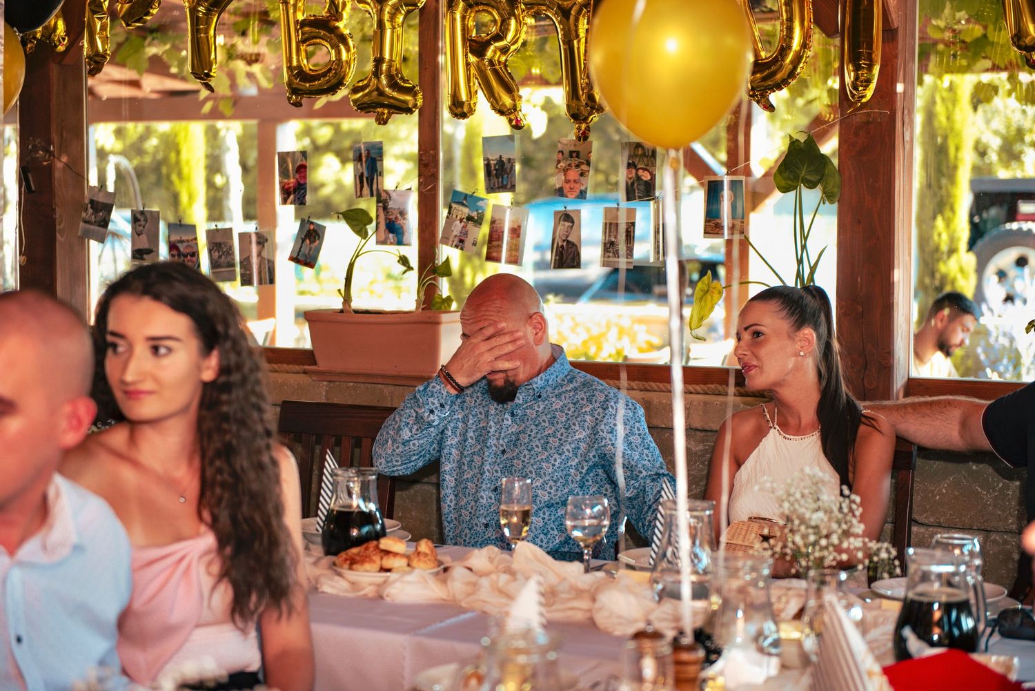 Emotional man covering his face in surprise at a birthday dinner table decorated with balloons and hanging photos.