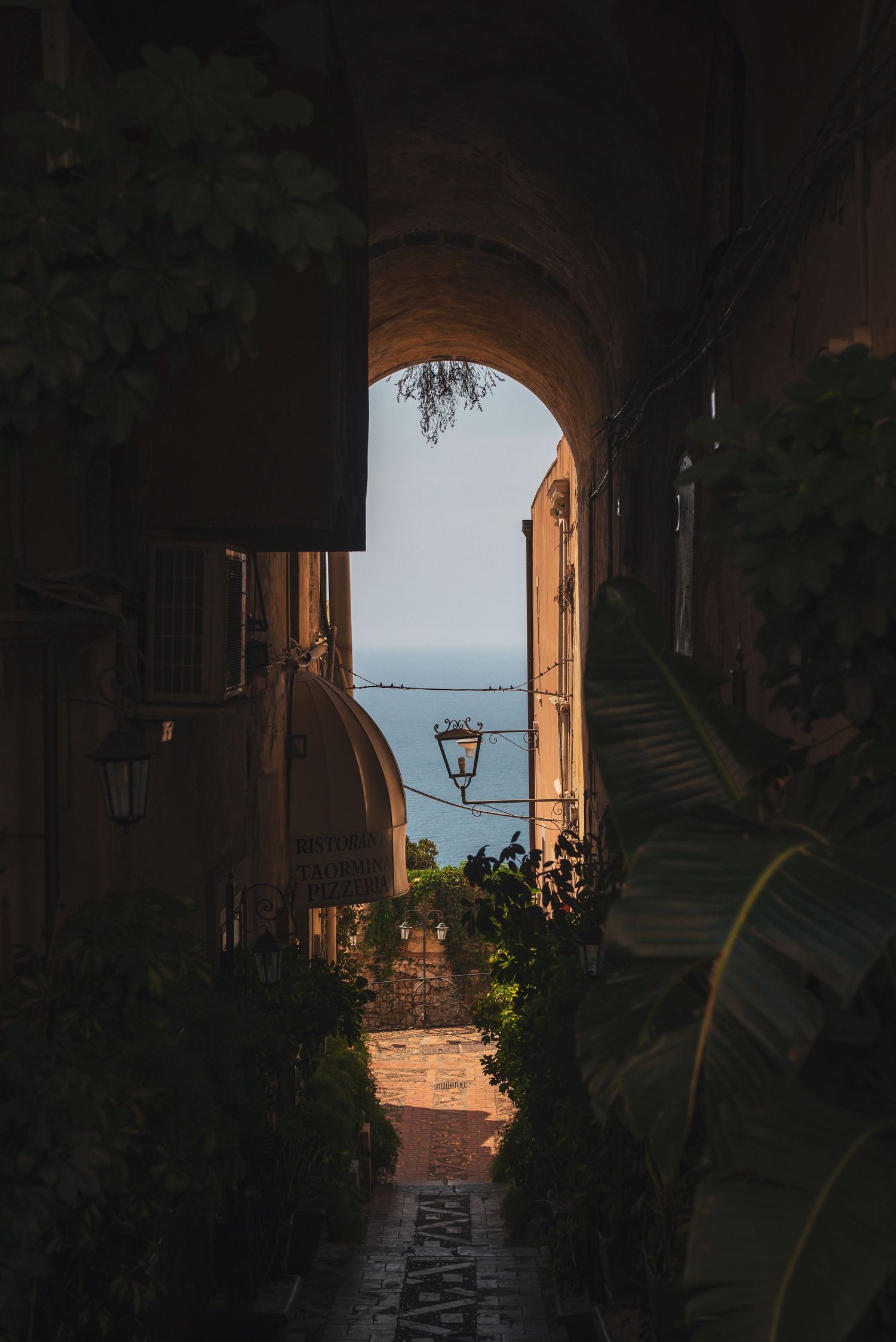 Dark, narrow stone alleyway in Taormina, Sicily, featuring an archway that frames a distant view of the sea