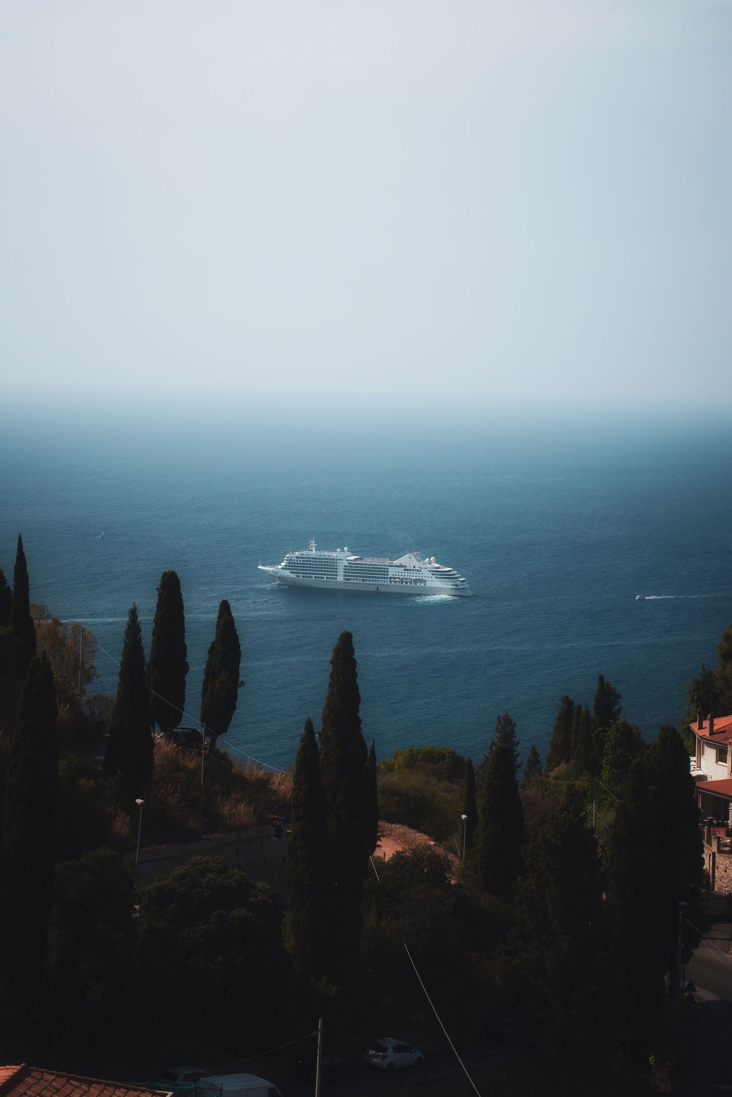 View of a cruise ship on the sea from the main public car park in Taormina, Sicily, framed by dark cypress trees on a hillside.