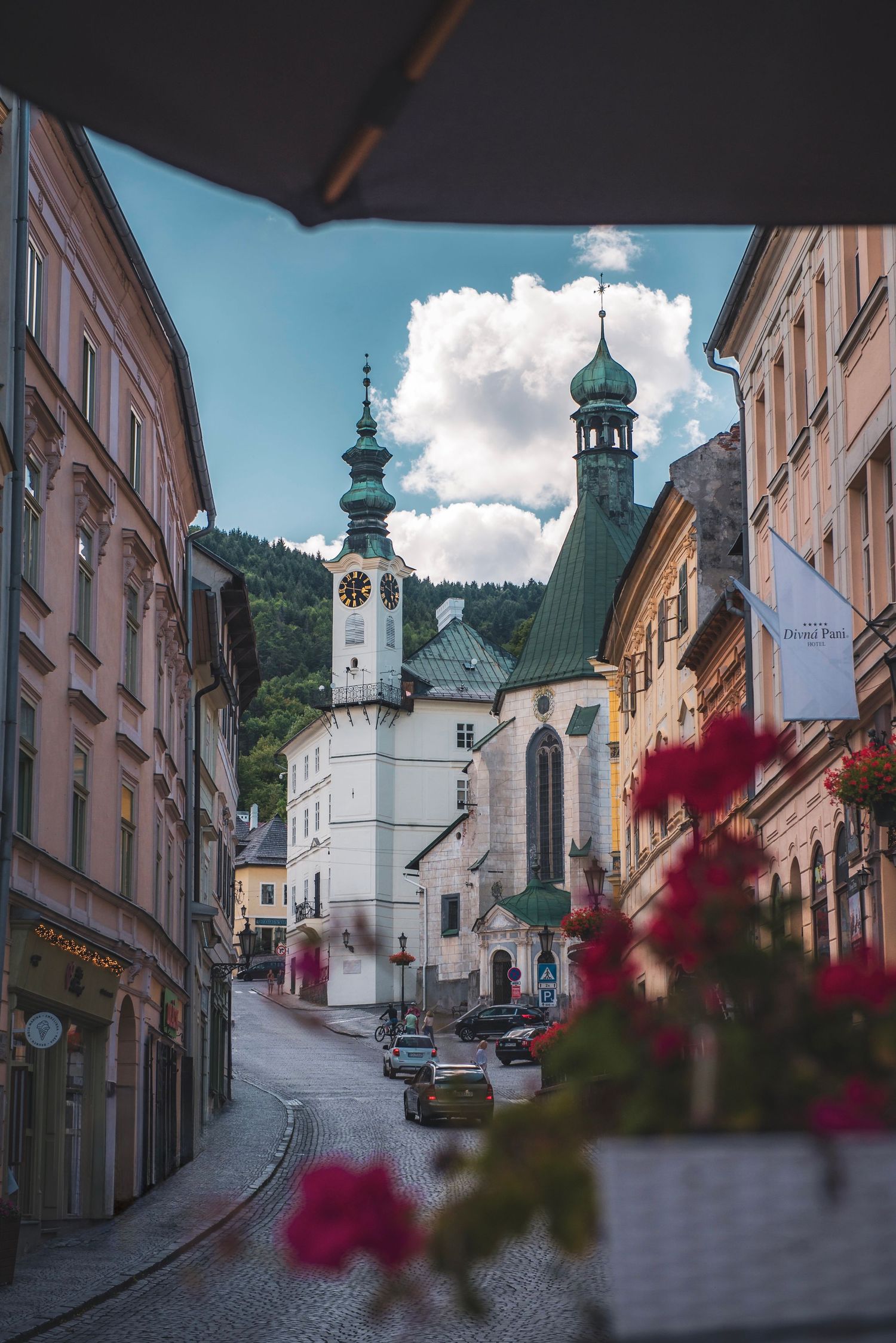 Street view in Banská Štiavnica, Slovakia, featuring the Town Hall tower and Church of St. Catherine framed by red flowers.