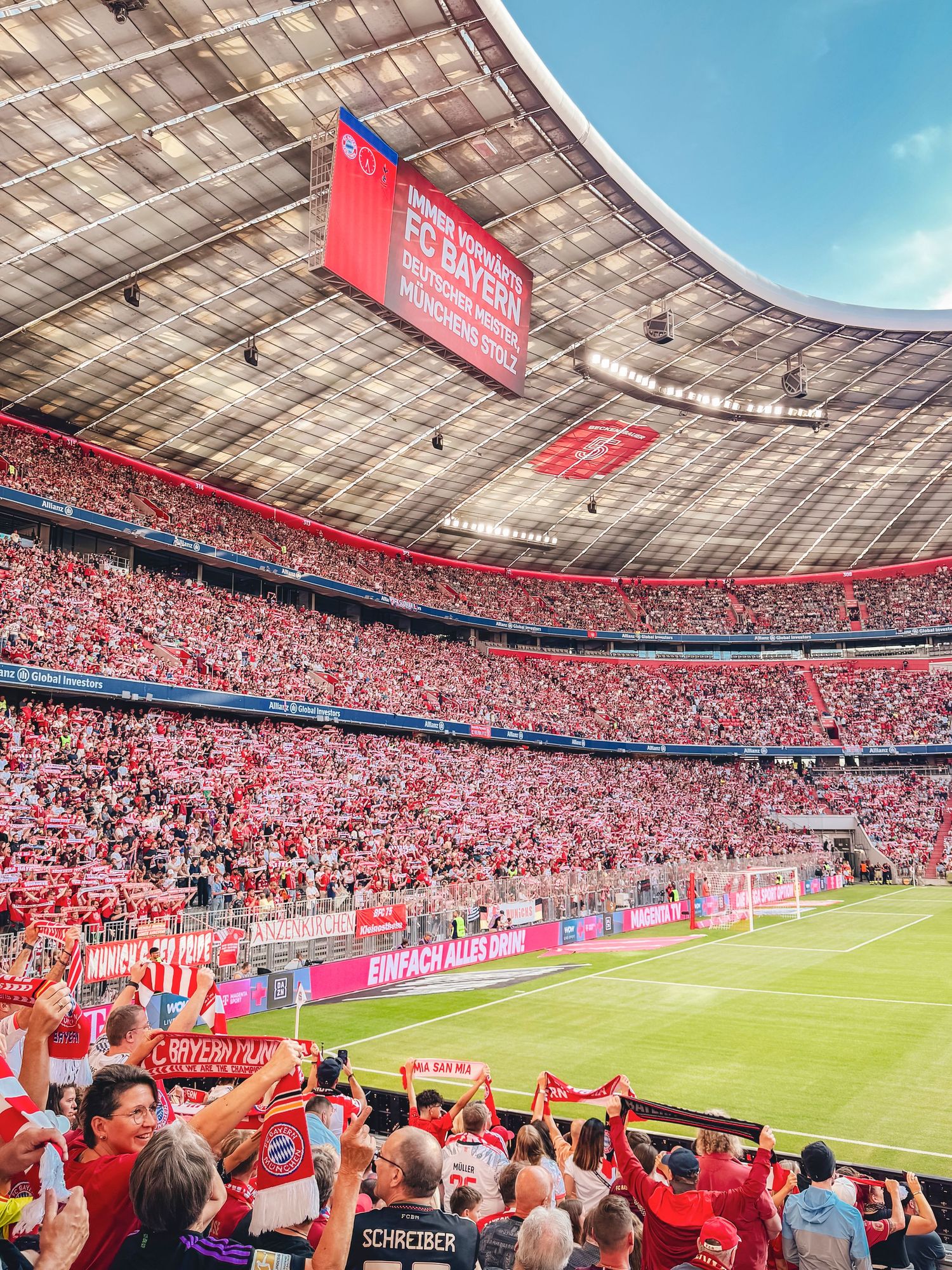 Packed stands at the Allianz Arena in Munich, Germany during a Bundesliga match, with fans cheering and holding Bayern Munich scarves.