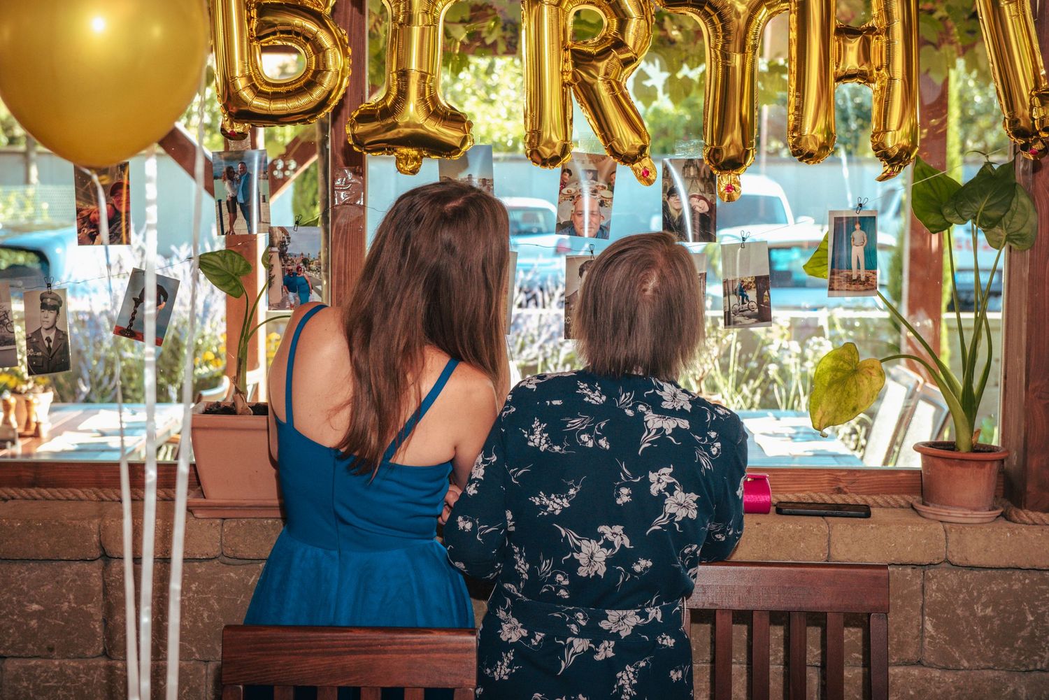 Two women looking at hanging photograph memories under gold balloons at a birthday event.