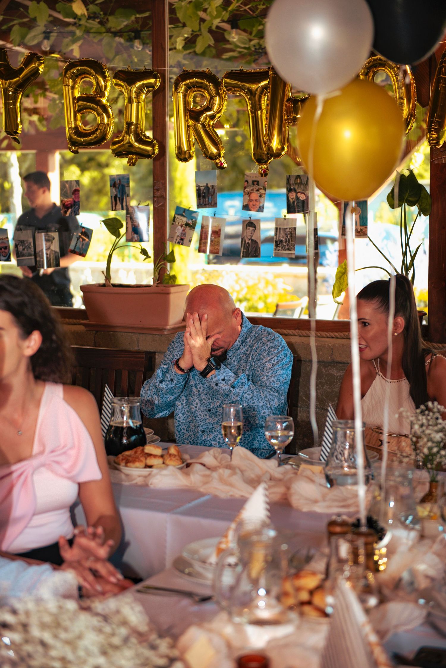 Emotional man covering his face in surprise at a birthday dinner table decorated with balloons and hanging photos.